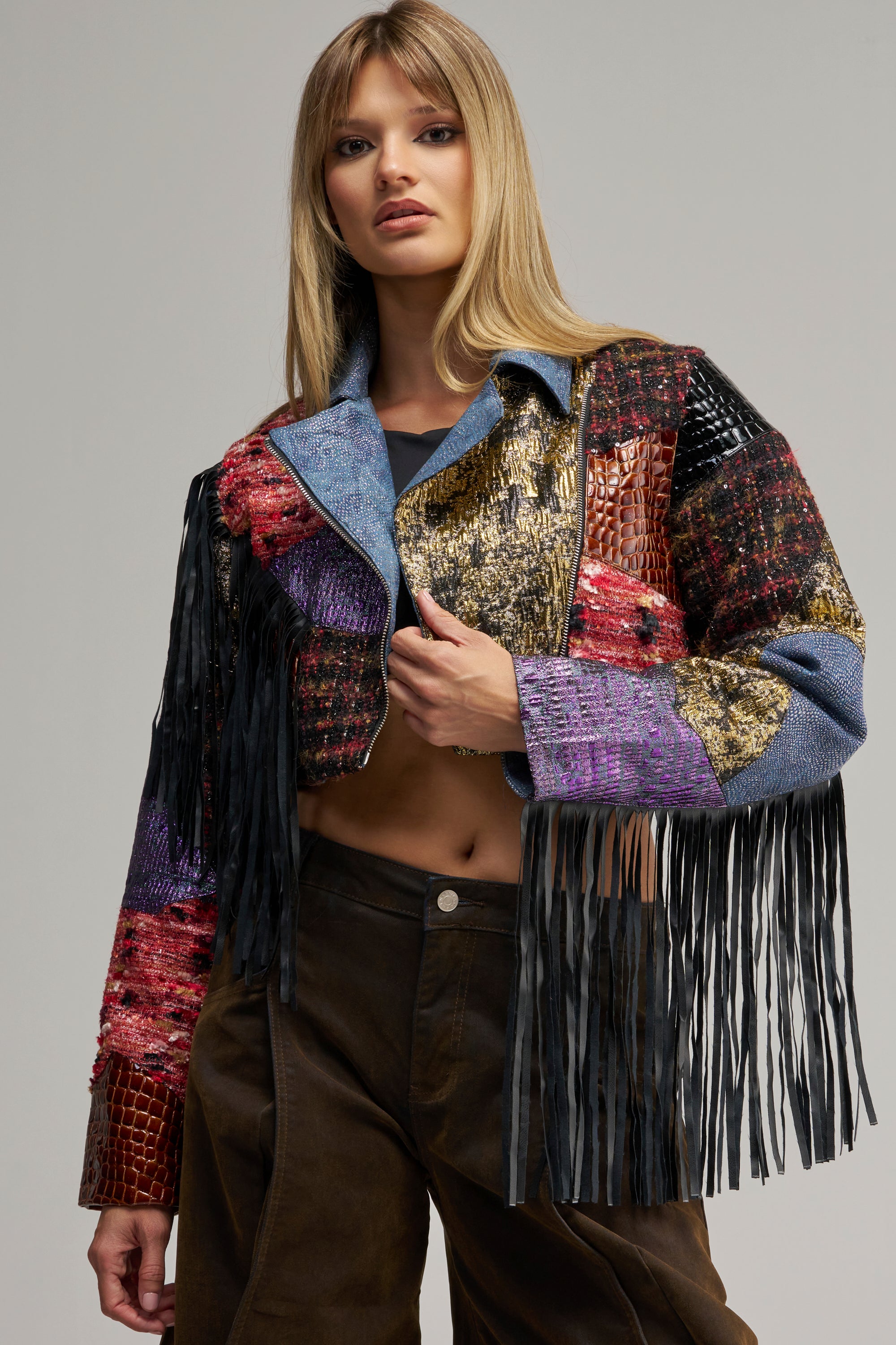 A woman with long blonde hair wears the CAN’T STOP WON’T STOP PATCHWORK MOTO, its vibrant patchwork design and long black fringe accentuating the sleeves. Paired with dark brown pants, she stands against a plain gray background, effortlessly channeling bold streetwear style.