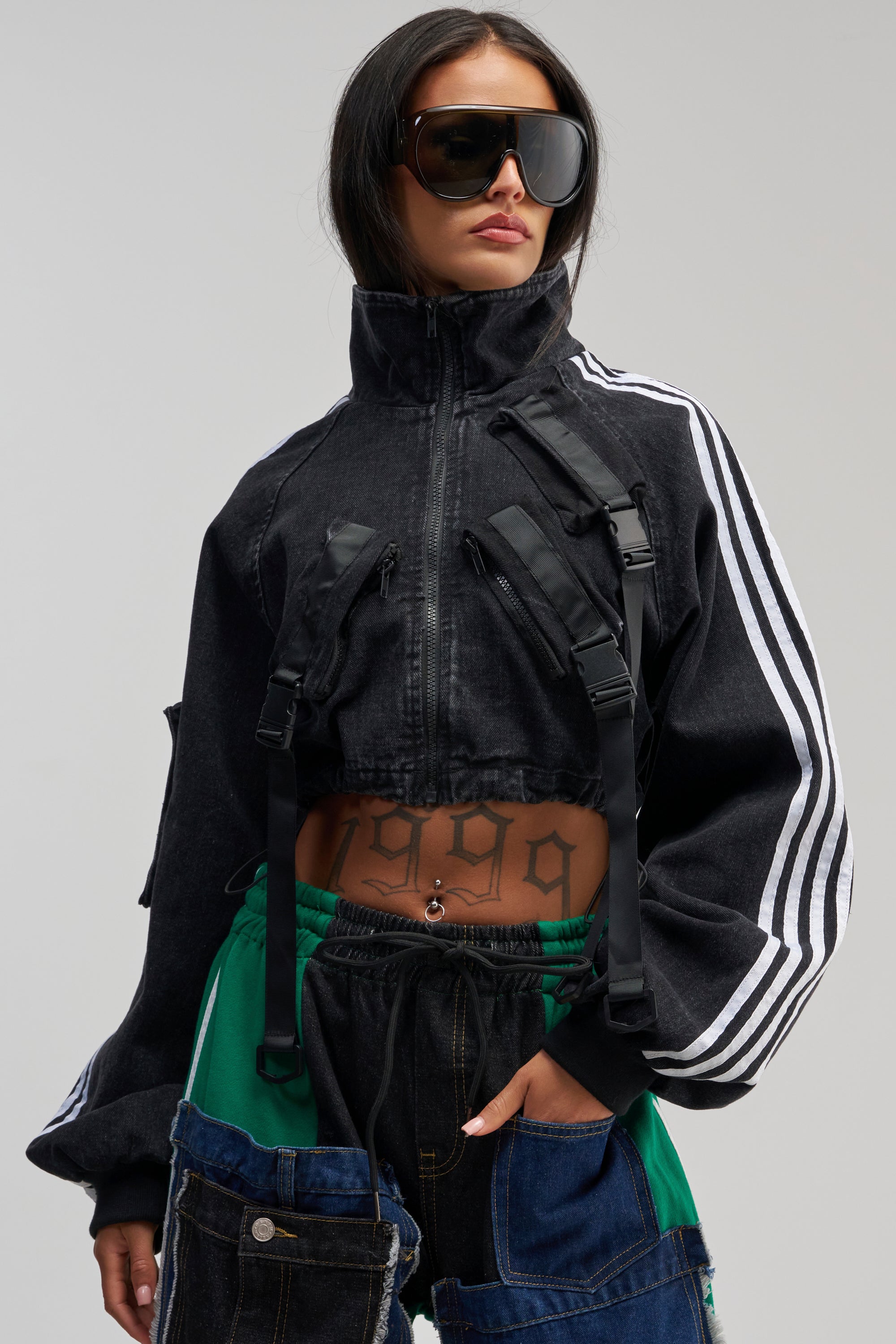 A woman wearing large sunglasses and the RAE CARGO DENIM JACKET with white stripes, paired with patchwork pants. She has a “1999” tattoo on her stomach and is posing with one hand in her pocket.