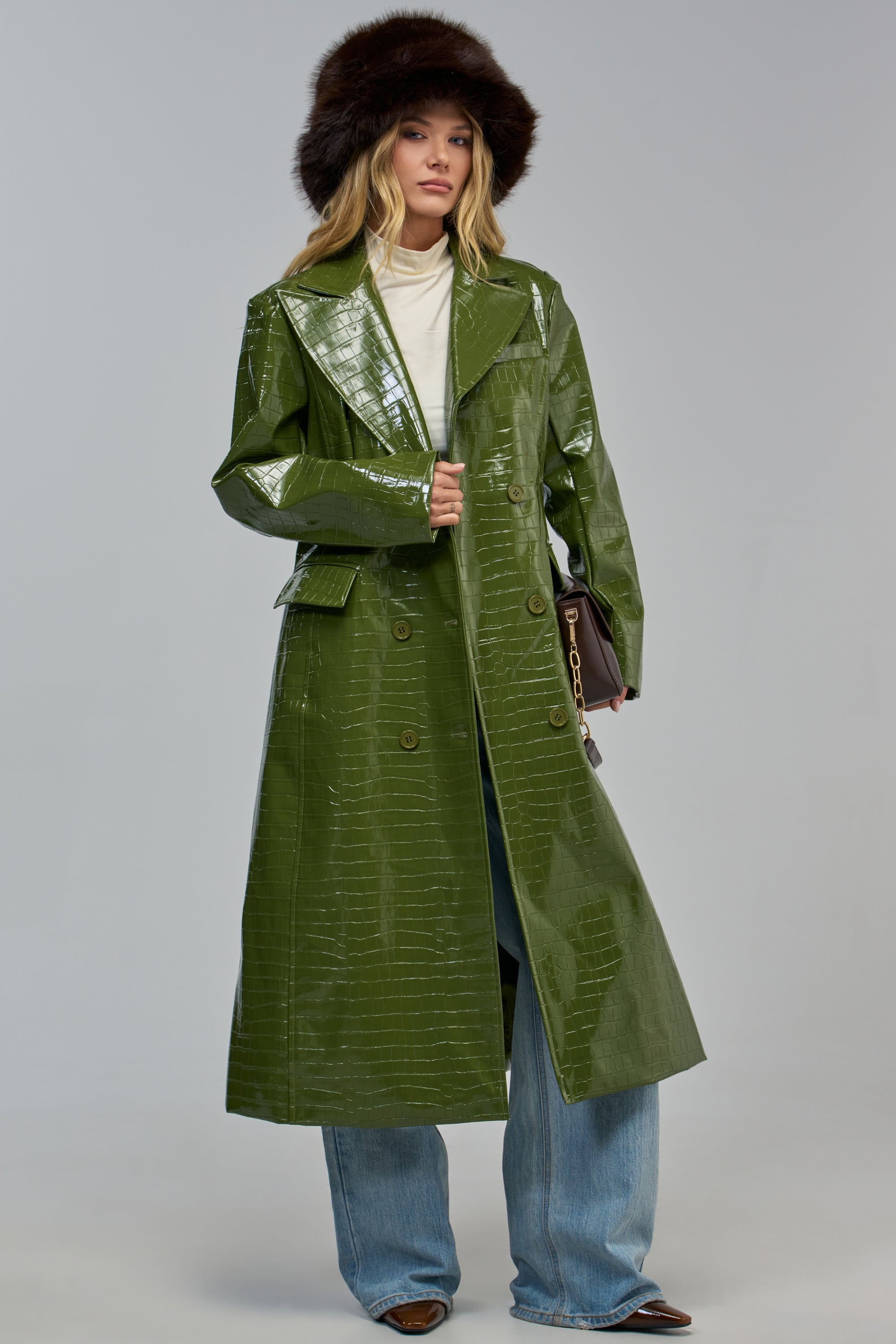 A person wears an olive IVY PATENT CROC TRENCH coat with a brown faux-fur hat and wide-leg jeans.