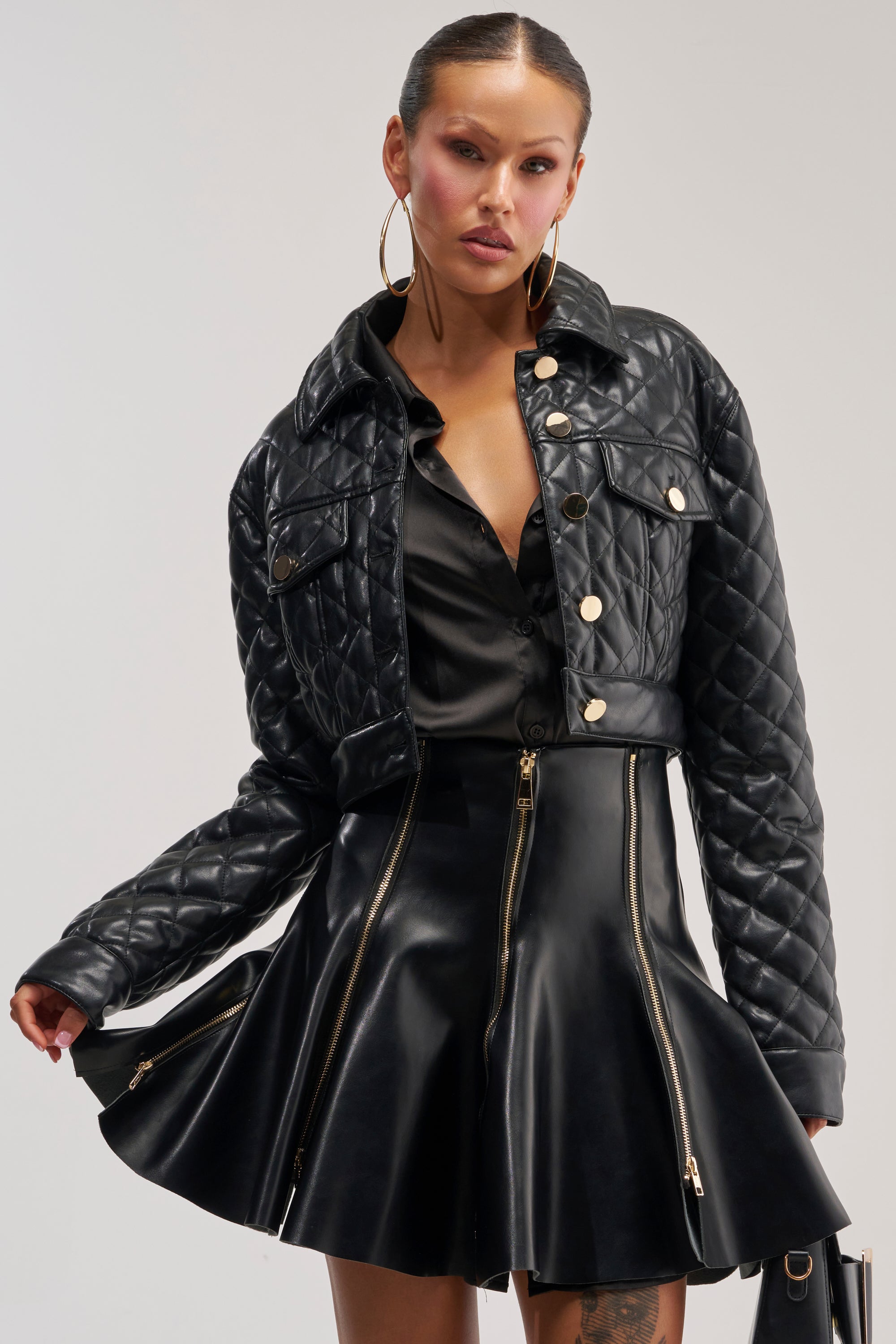 A woman poses confidently in the ROMY QUILTED FAUX LEATHER JACKET, paired with a flared black leather skirt featuring gold-tone hardware. She holds the sides of her skirt, accessorized with hoop earrings and slicked-back hair.