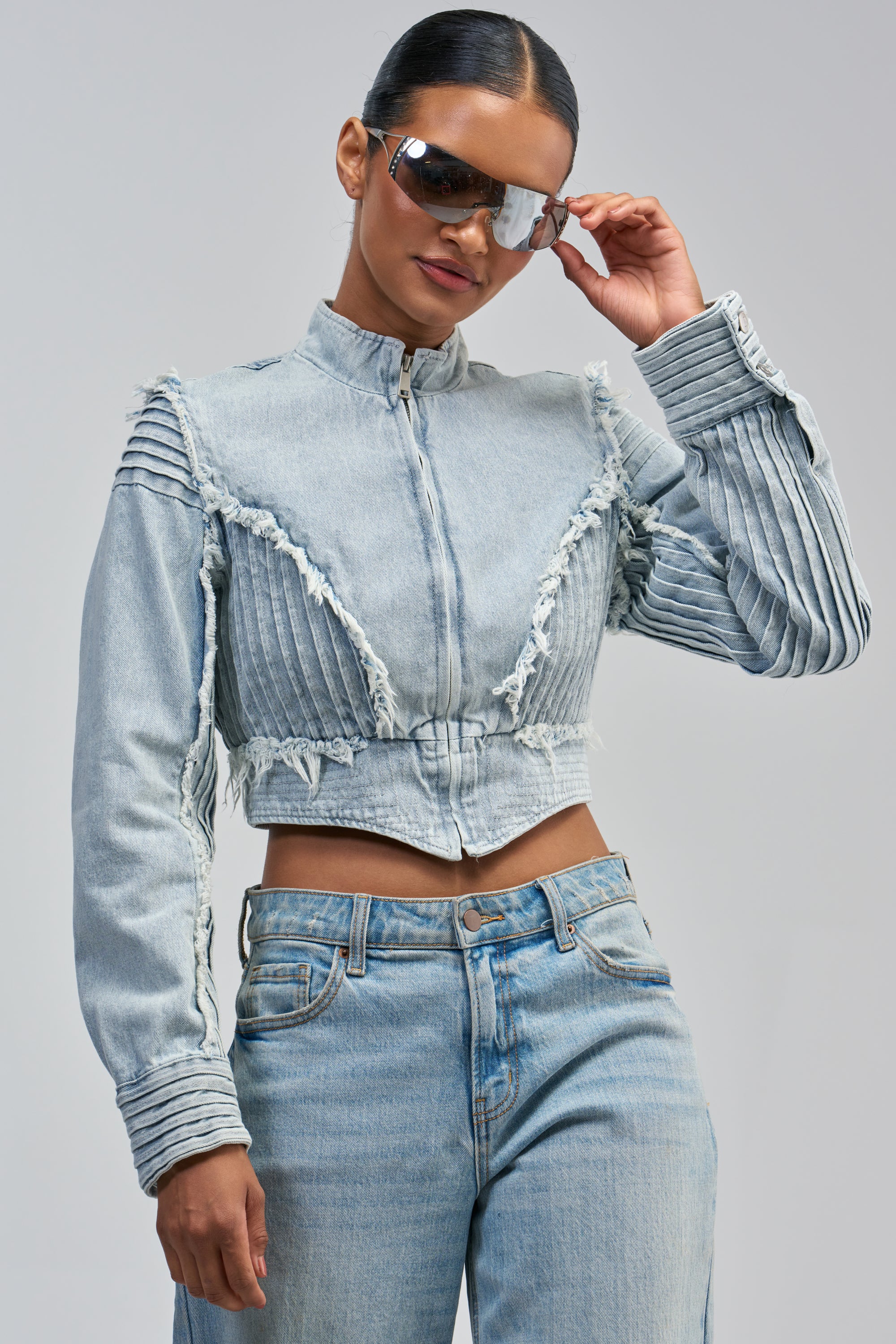 A woman wearing large sunglasses and the IRIS DENIM MOTO—a light blue cropped moto jacket with pleated and frayed details—paired with light blue jeans stands against a plain background.
