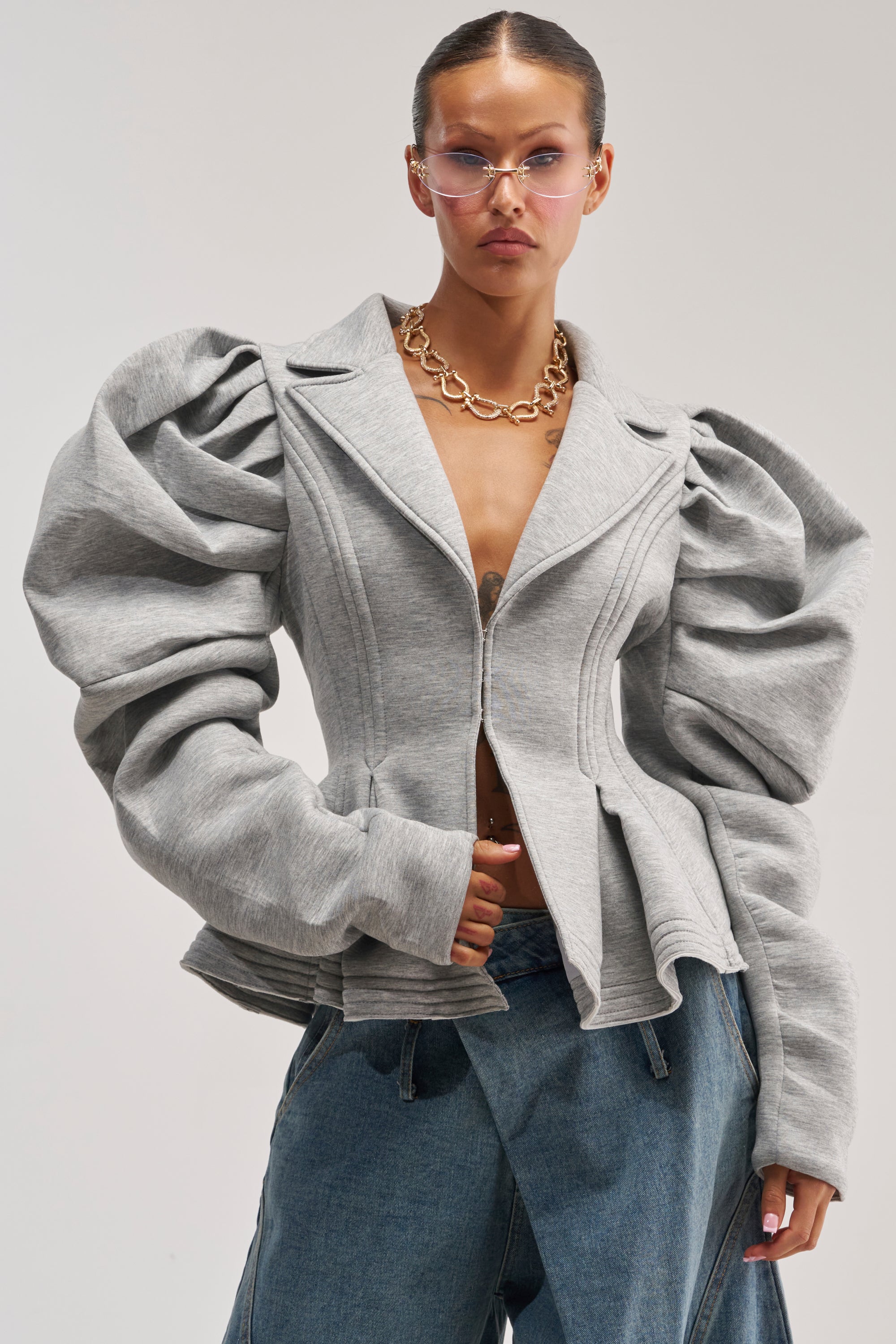 A person wearing the LIORA PEPLUM BLAZER in gray with exaggerated puff sleeves, paired with blue jeans, a chunky gold chain necklace, and pink-tinted sunglasses, poses confidently against a plain background.