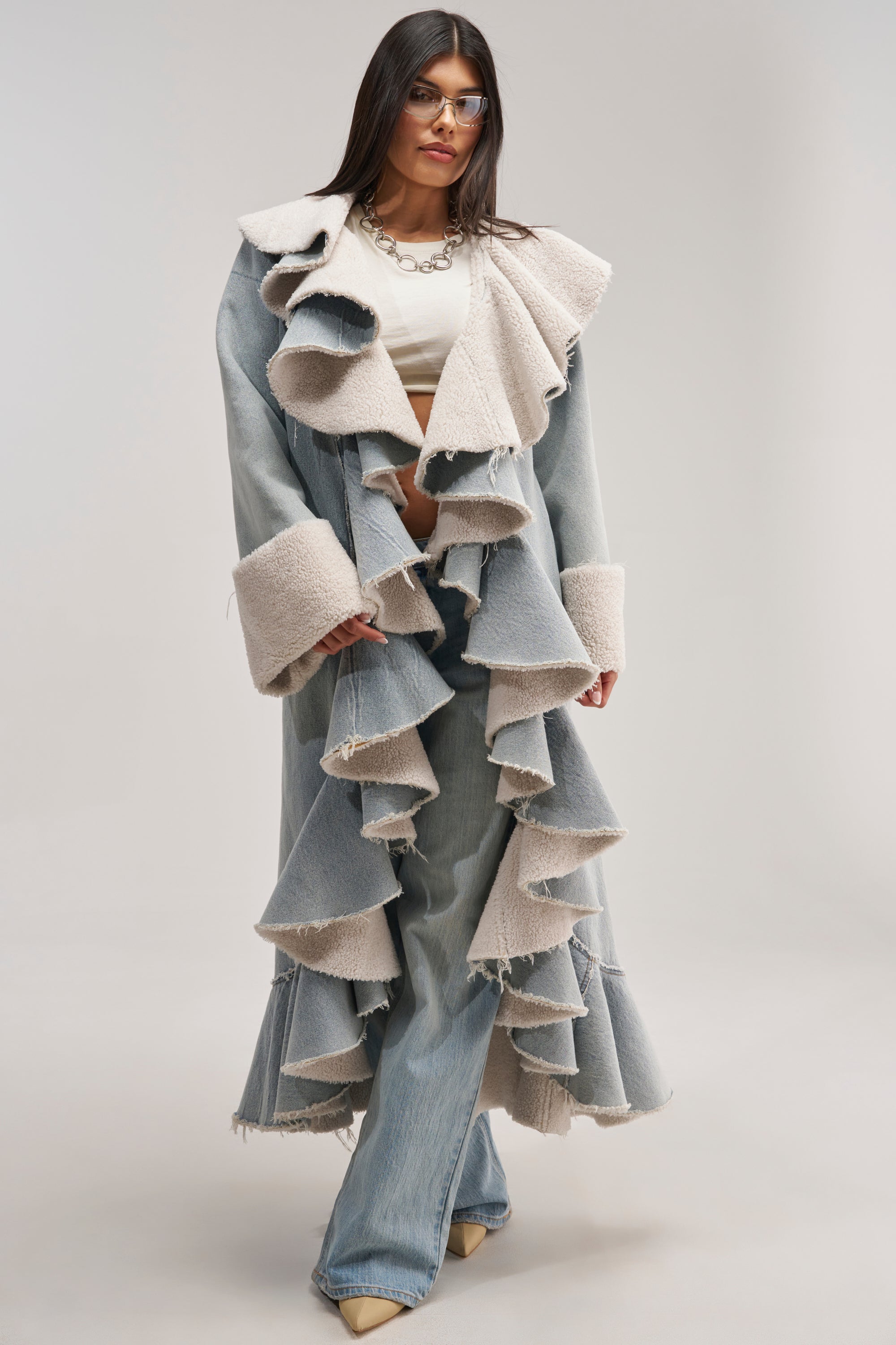 Wearing the AIN'T ABOUT THE MONEY DENIM TRENCH, a woman in glasses stands against a plain light background. The trench features dramatic ruffles, beige fleece trim, and an oversized shawl collar, which she pairs with beige pointed shoes.