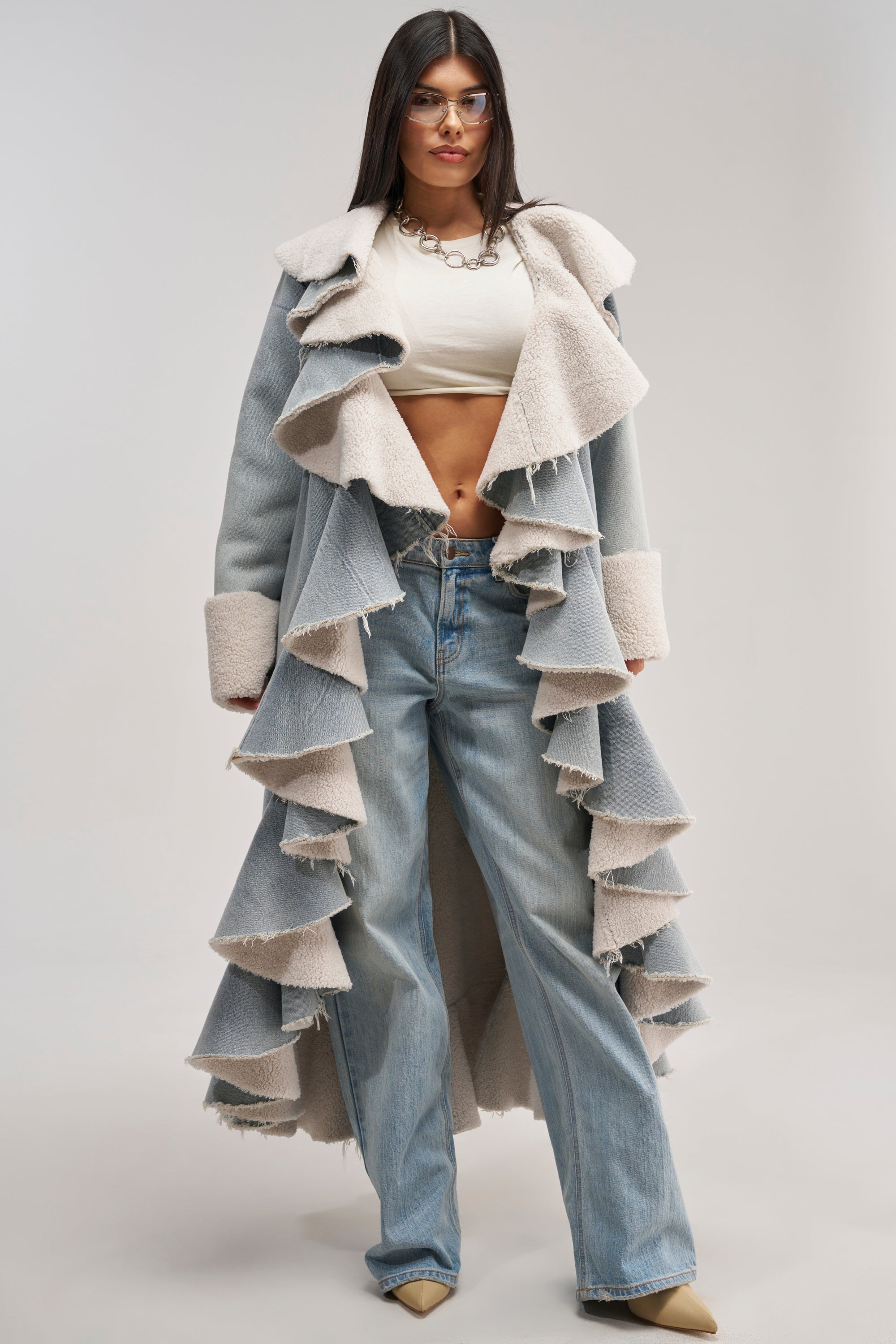 A person wearing glasses, a white cropped top, loose light blue jeans, and beige pointed shoes stands facing forward against a plain background, showcasing the AIN'T ABOUT THE MONEY DENIM TRENCH—a long ruffled light blue and beige sherpa trench with an oversized shawl collar and faux sherpa lining.