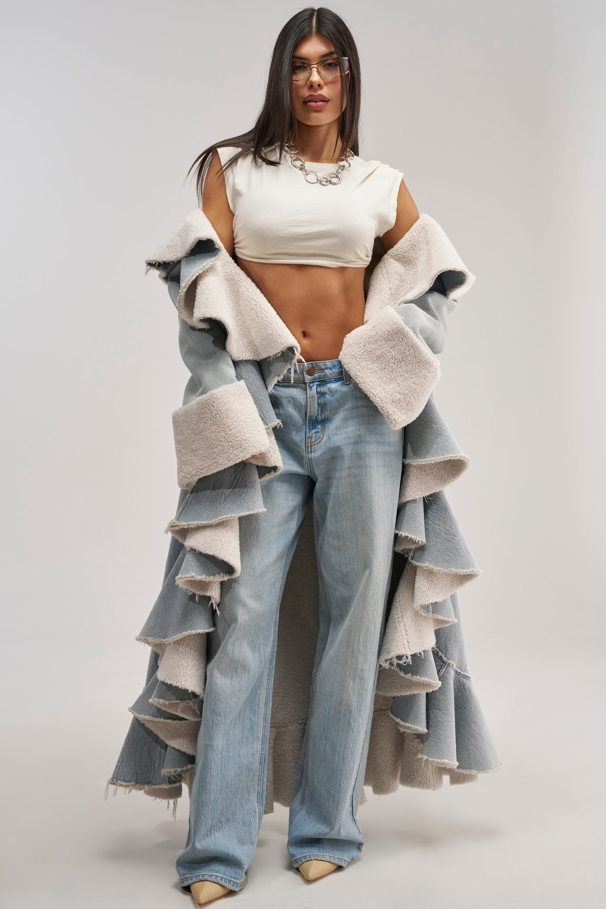 A woman wearing glasses, a white crop top, light blue wide-leg jeans, and the AIN'T ABOUT THE MONEY DENIM TRENCH—with its oversized shawl collar and faux sherpa lining—stands confidently against a plain background.
