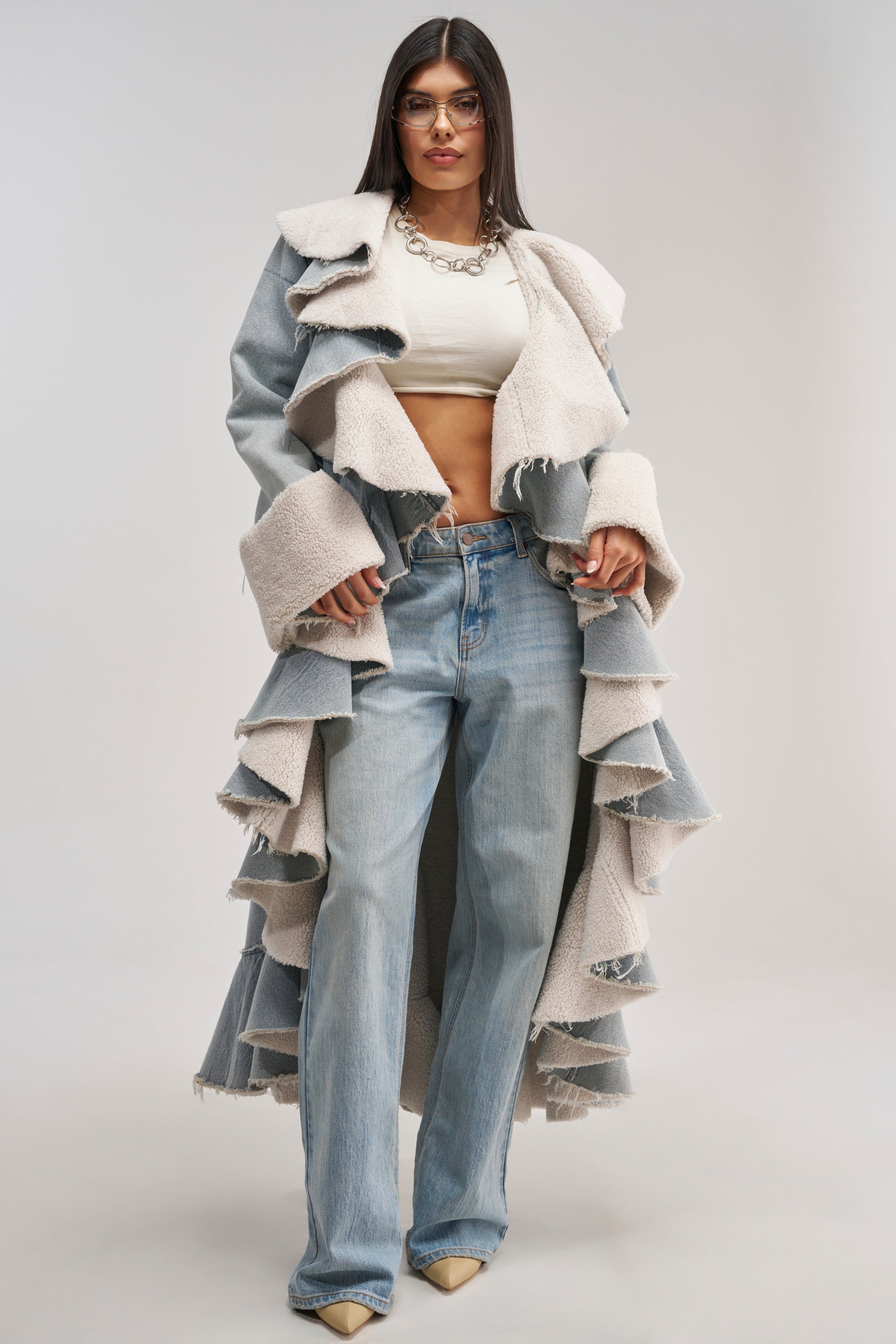 Wearing the AIN'T ABOUT THE MONEY DENIM TRENCH—featuring ruffled details and shearling accents—a woman in glasses stands against a plain light background, layering the longline denim coat over a white crop top, loose blue jeans, and beige heels.