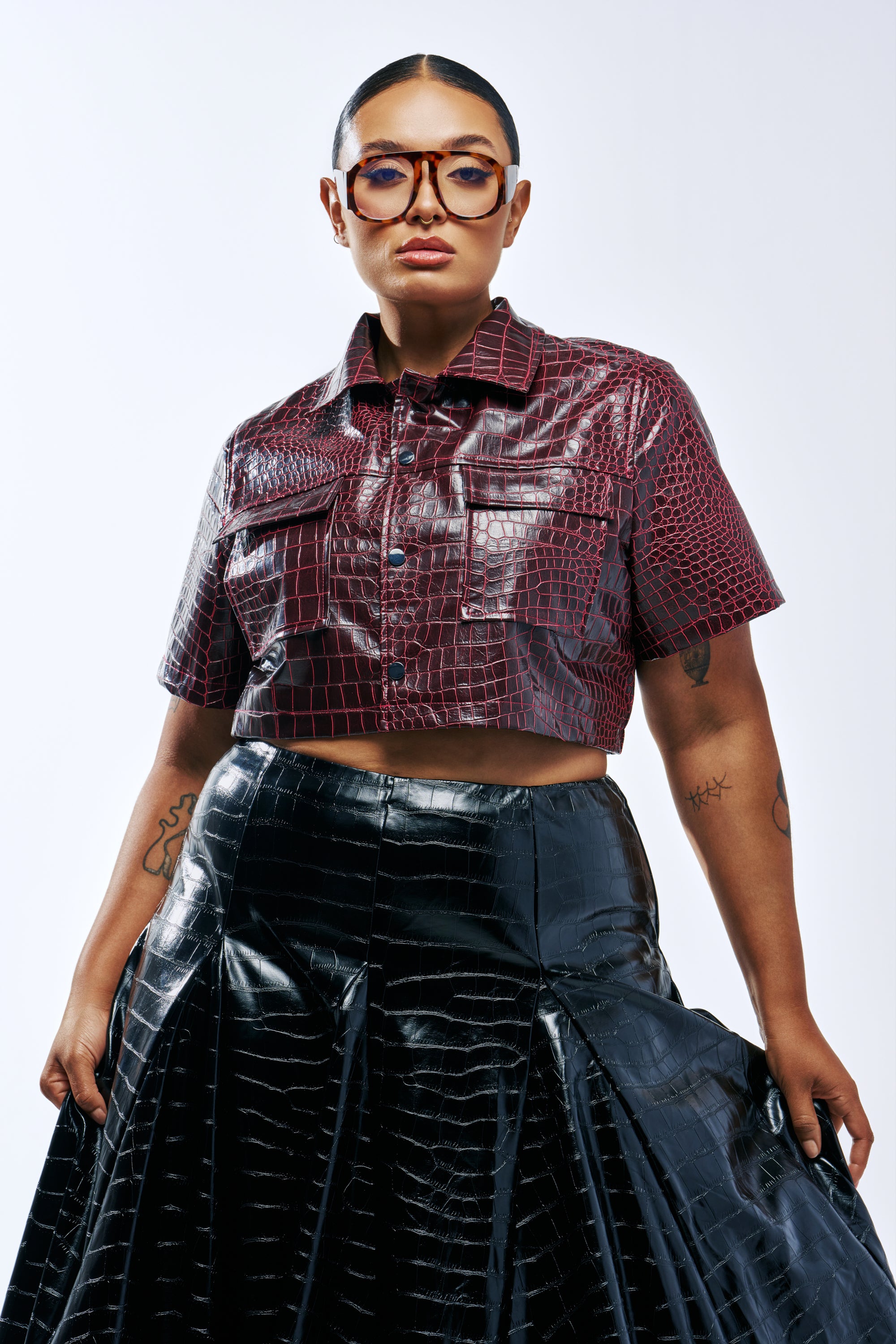 A person wearing a BORDEAUX CROC UTILITY CROP BUTTON DOWN in burgundy faux leather paired with a black crocodile-embossed pleated skirt.