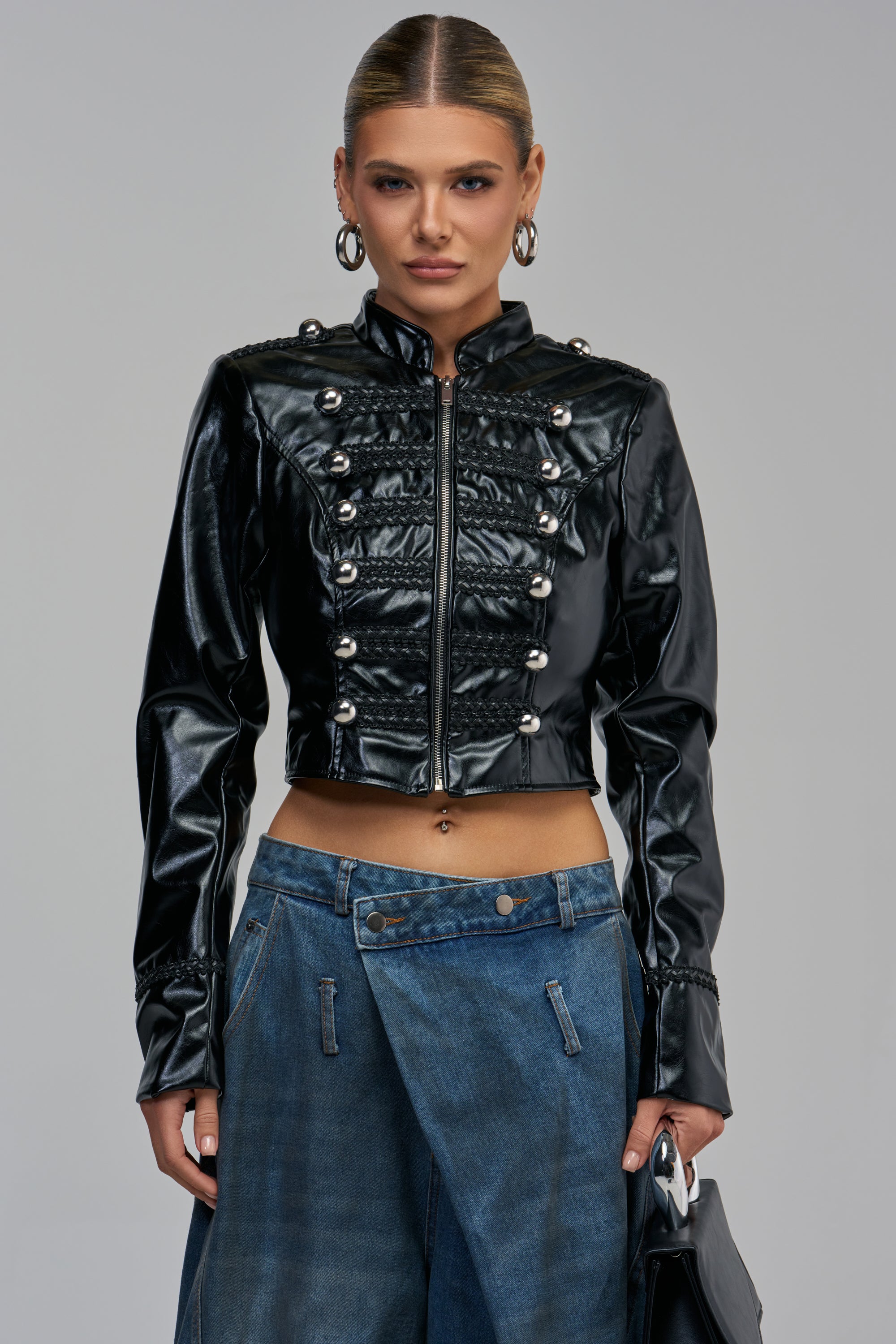 Model wearing THEE BADDEST PU MOTO, a cropped black faux leather military moto jacket with silver button details and braided trim.