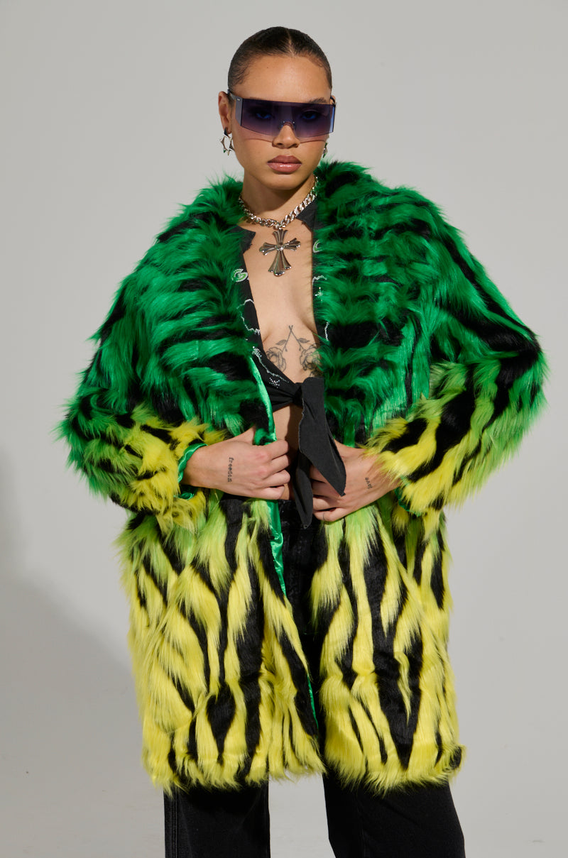 A person wearing the SOUND THE ALARM FAUX FUR COAT in vibrant green and yellow stands confidently in their zebra-striped shoes. They accessorize with dark sunglasses, a necklace, and display a tattoo on their chest, set against a plain, neutral background.
