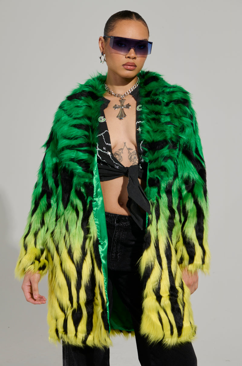 A person confidently stands wearing the SOUND THE ALARM FAUX FUR COAT, featuring bold green and yellow ombre patterns. They have short, tied-back hair, zebra-striped sunglasses, a black top, and a chain necklace with a cross pendant against a plain background.