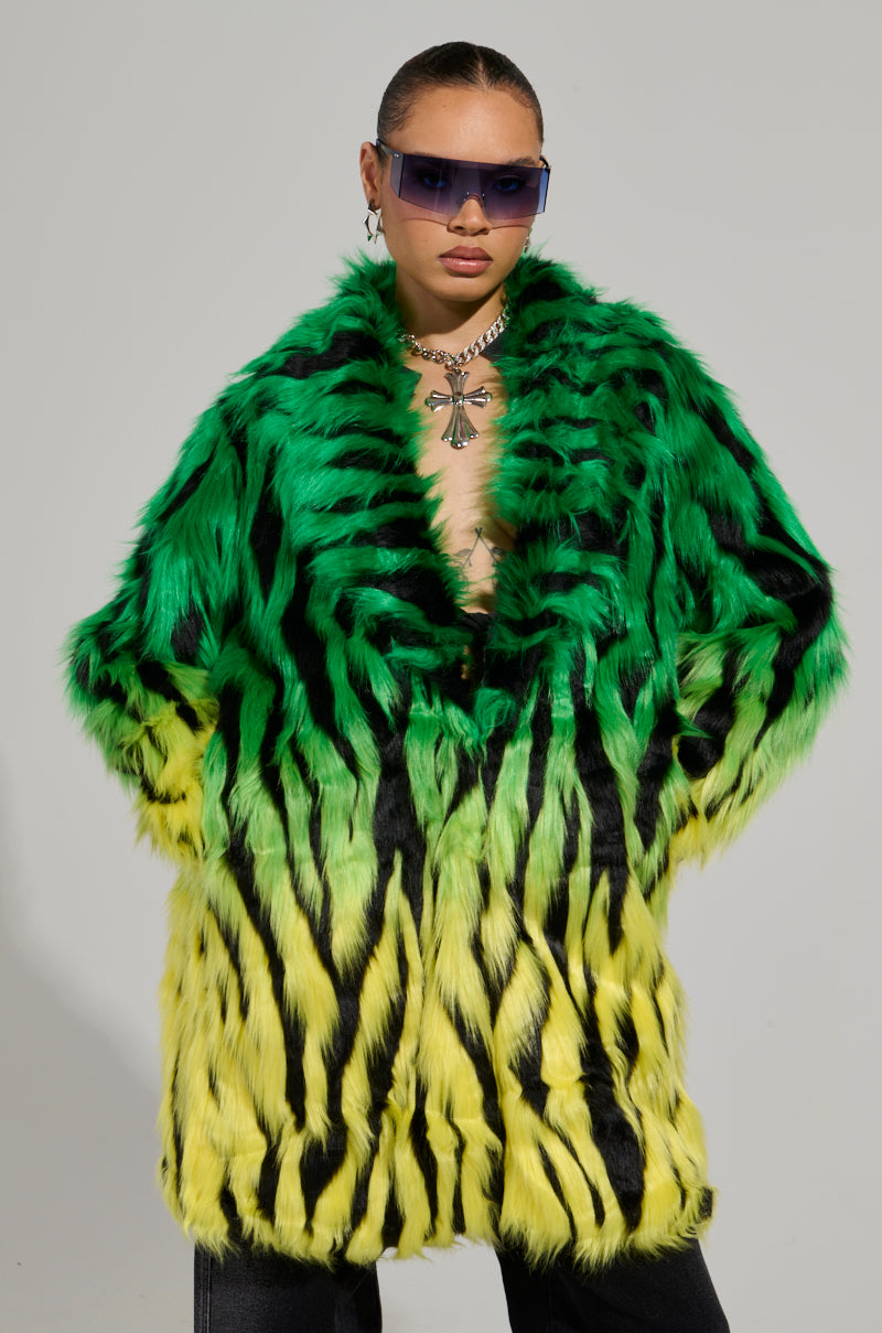 Wearing the SOUND THE ALARM FAUX FUR COAT, characterized by its vibrant neon green and yellow stripes, a person with short hair complements the look with dark sunglasses and a chunky necklace against a plain gray backdrop.