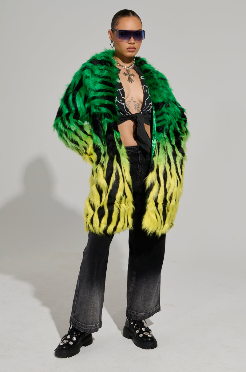 Clad in the SOUND THE ALARM FAUX FUR COAT with its striking green and yellow neon colorway, and paired with a black top, dark jeans, chunky black shoes, sunglasses, and tattoos visible, they exude confidence against a plain backdrop.