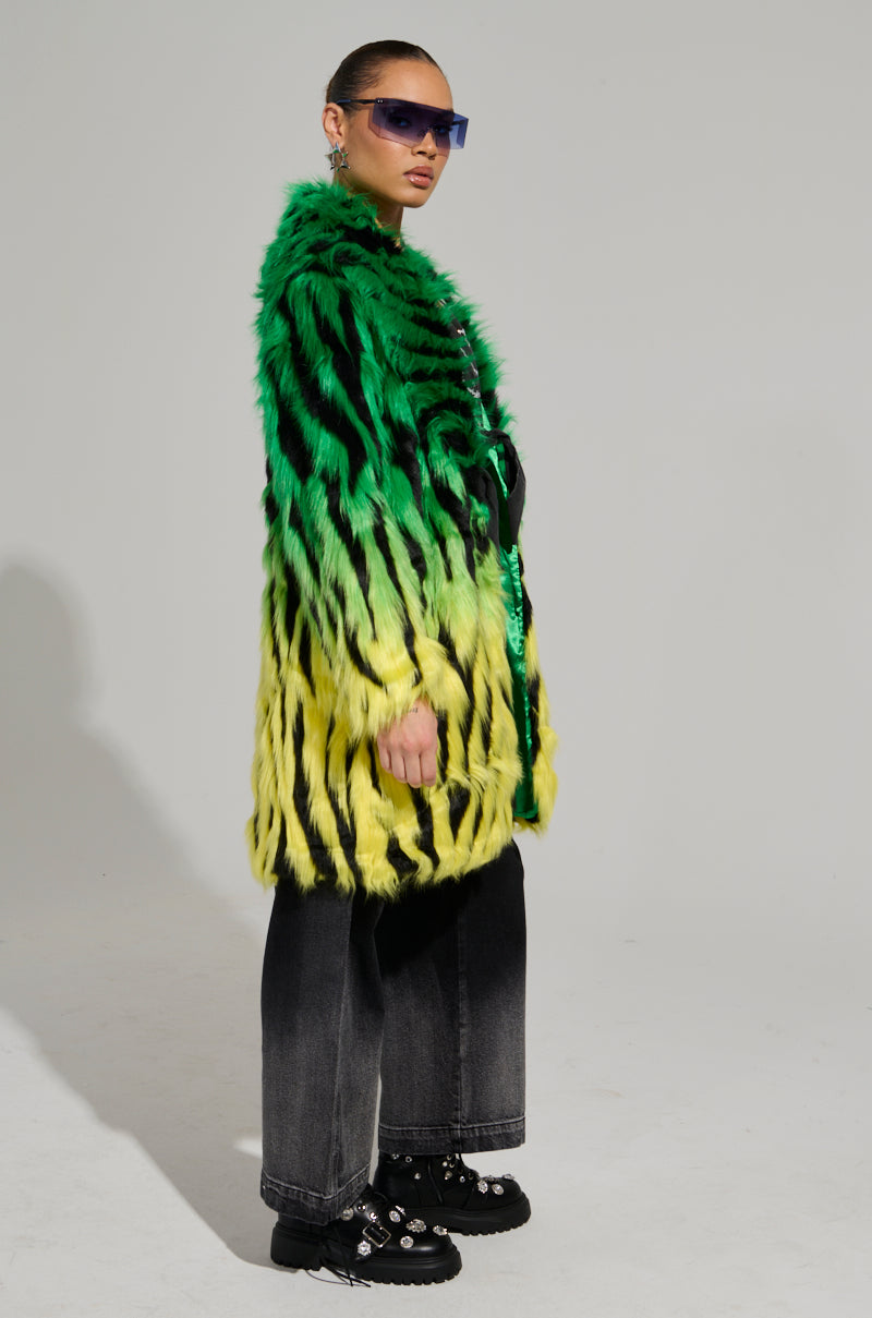 Someone confidently dons the SOUND THE ALARM FAUX FUR COAT, which features a vibrant zebra-striped design. Their hair is slicked back, and they accessorize with blue-tinted sunglasses. Completing the look are black wide-leg jeans and embellished black shoes. A plain gray background allows their neon-colored outfit to stand out even more.