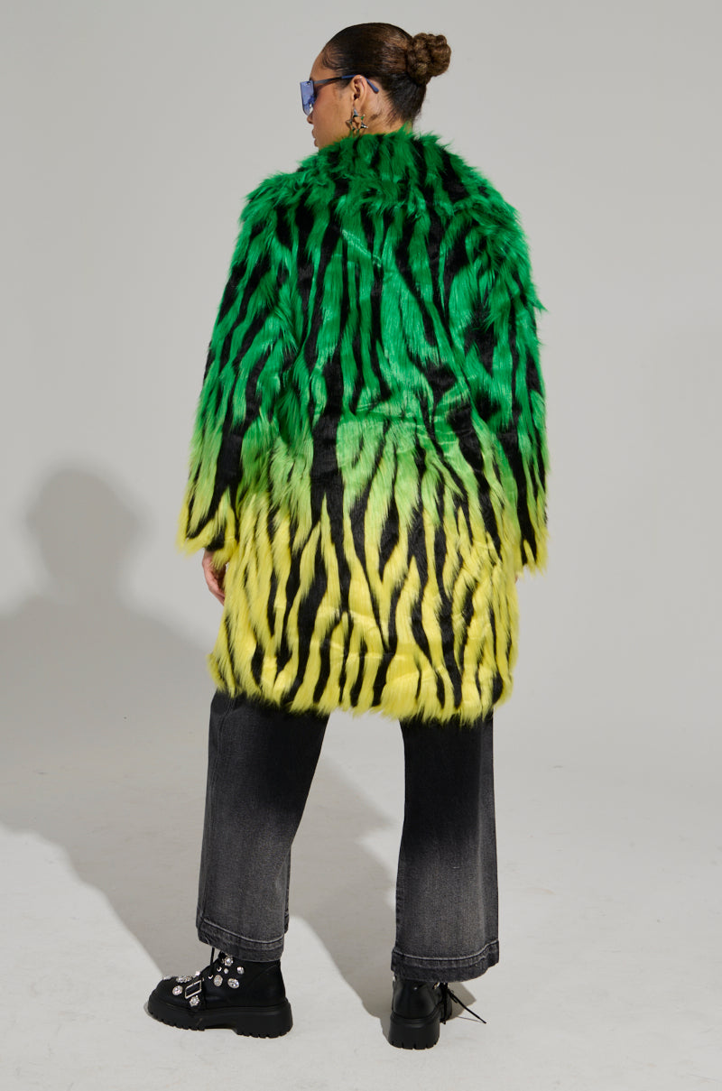 A person wearing the SOUND THE ALARM FAUX FUR COAT, a vibrant green and yellow zebra-striped design, paired with dark jeans and black boots. They sport sunglasses and have their hair styled in a bun, standing against a plain background.