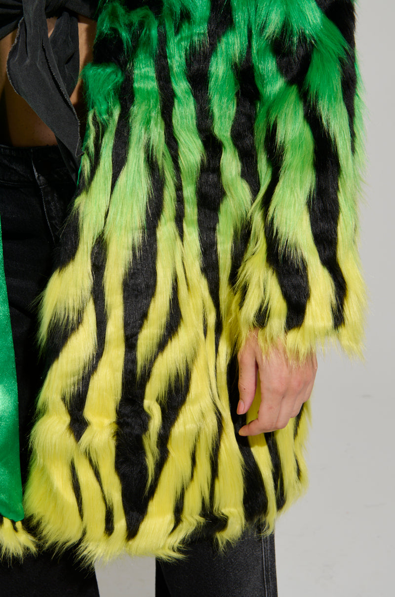 Close-up of a person wearing the SOUND THE ALARM FAUX FUR COAT, featuring a vibrant neon green, yellow, and black tiger stripe pattern. The eye-catching zebra-striped design offers a fluffy texture and is complemented by dark pants. The person's hand is visibly hanging at their side.