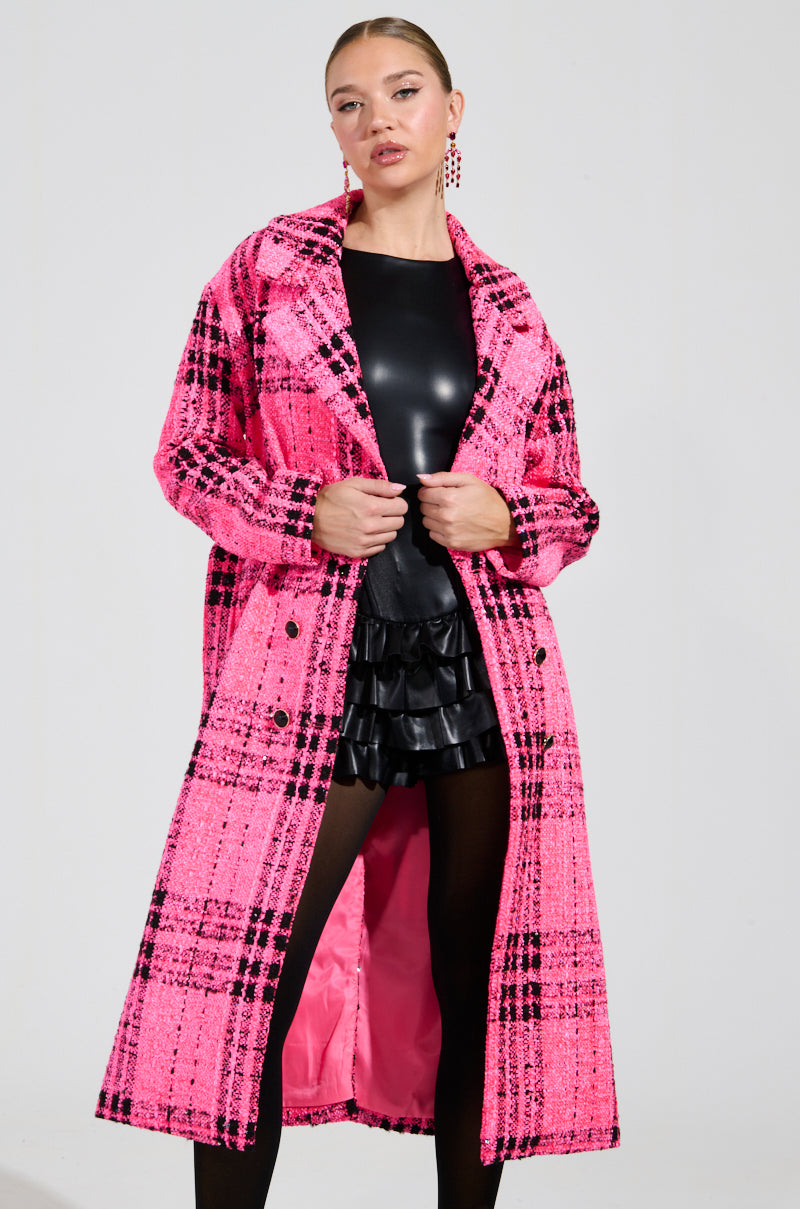 The individual poses in a SAINT JAMES TWEED TRENCH, featuring a long pink plaid design, layered over a sleek black leather ensemble. The open coat showcases a ruffled skirt beneath. Their hair is styled in a slicked-back fashion against a plain backdrop.