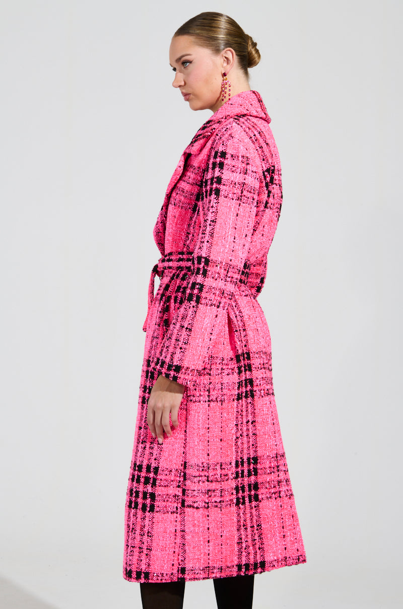A person with tied-back hair is wearing the SAINT JAMES TWEED TRENCH, a long pink plaid coat featuring a tie belt, and they are standing sideways against a plain white background.