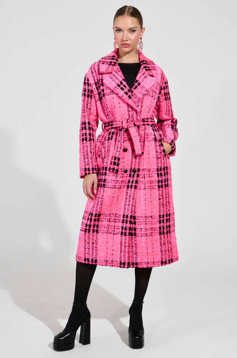 Dressed in the vibrant SAINT JAMES TWEED TRENCH, featuring a bold pink plaid pattern and a chic belted waist, a person stands confidently. They complement the trench coat with black tights and chunky-heeled shoes against a plain background that highlights the stylishness of their outfit.