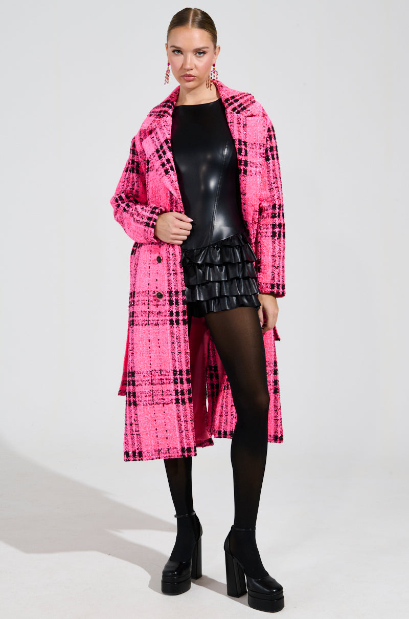 A model poses confidently in the SAINT JAMES TWEED TRENCH, featuring a vibrant pink plaid pattern. She pairs it with a sleek black leather top and ruffled skirt, complemented by black tights and platform shoes. Her hair is pulled back, showcasing her statement earrings against a plain white background.
