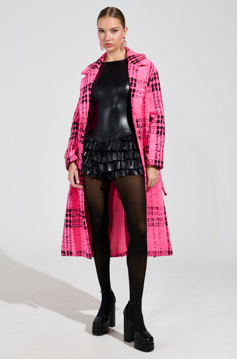 A person is wearing the SAINT JAMES TWEED TRENCH, styled over a black leather-look dress featuring ruffled details and subtle sequin embellishments. They pair this ensemble with black tights and platform shoes, standing confidently against a plain white background.