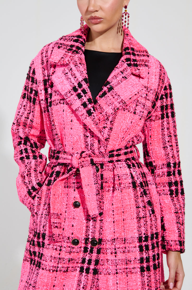 A person wearing the SAINT JAMES TWEED TRENCH in a vibrant pink plaid, with a belt elegantly cinched at the waist. The coat features a sophisticated double-breasted design and is beautifully complemented by matching pink earrings, creating a chic ensemble that stands out against the plain, light-colored background.