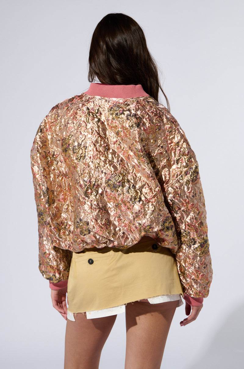 A person with long brown hair is shown from the back, wearing a shiny, textured OLLARI BROCADE BOMBER jacket featuring a pink collar and cuffs. They are also dressed in a light brown skirt with black buttons and a white layer underneath. The background is plain and light gray.