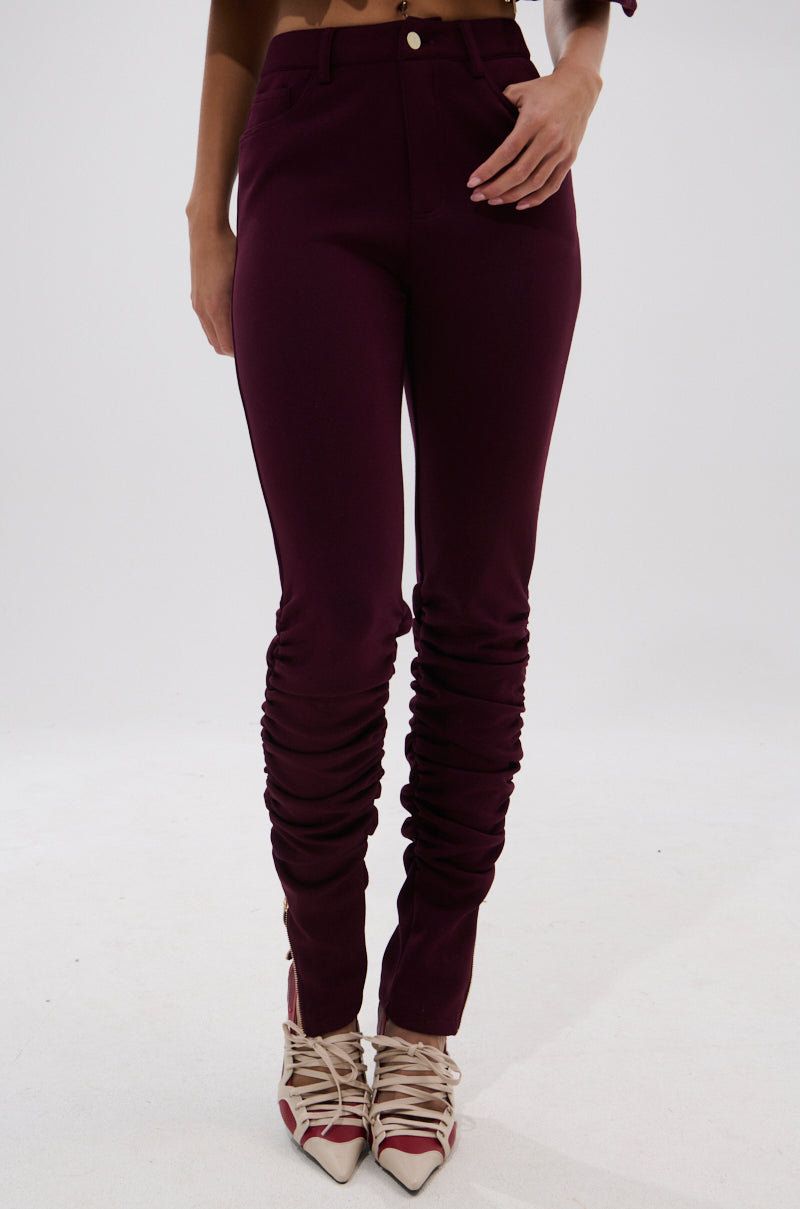 A person wears LYNX STACKED TROUSER IN BURGUNDY with ruched lower legs, paired with pointed-toe beige heels featuring crisscross ankle straps. The background is plain white. 2