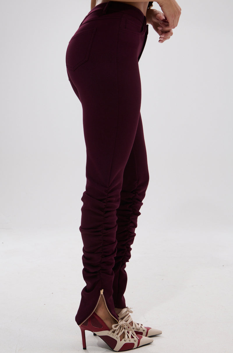 A person is shown from the waist down wearing fitted LYNX STACKED TROUSER IN BURGUNDY with a ruched finish and pointed high-heeled shoes with beige and burgundy accents, standing against a plain white background. 3