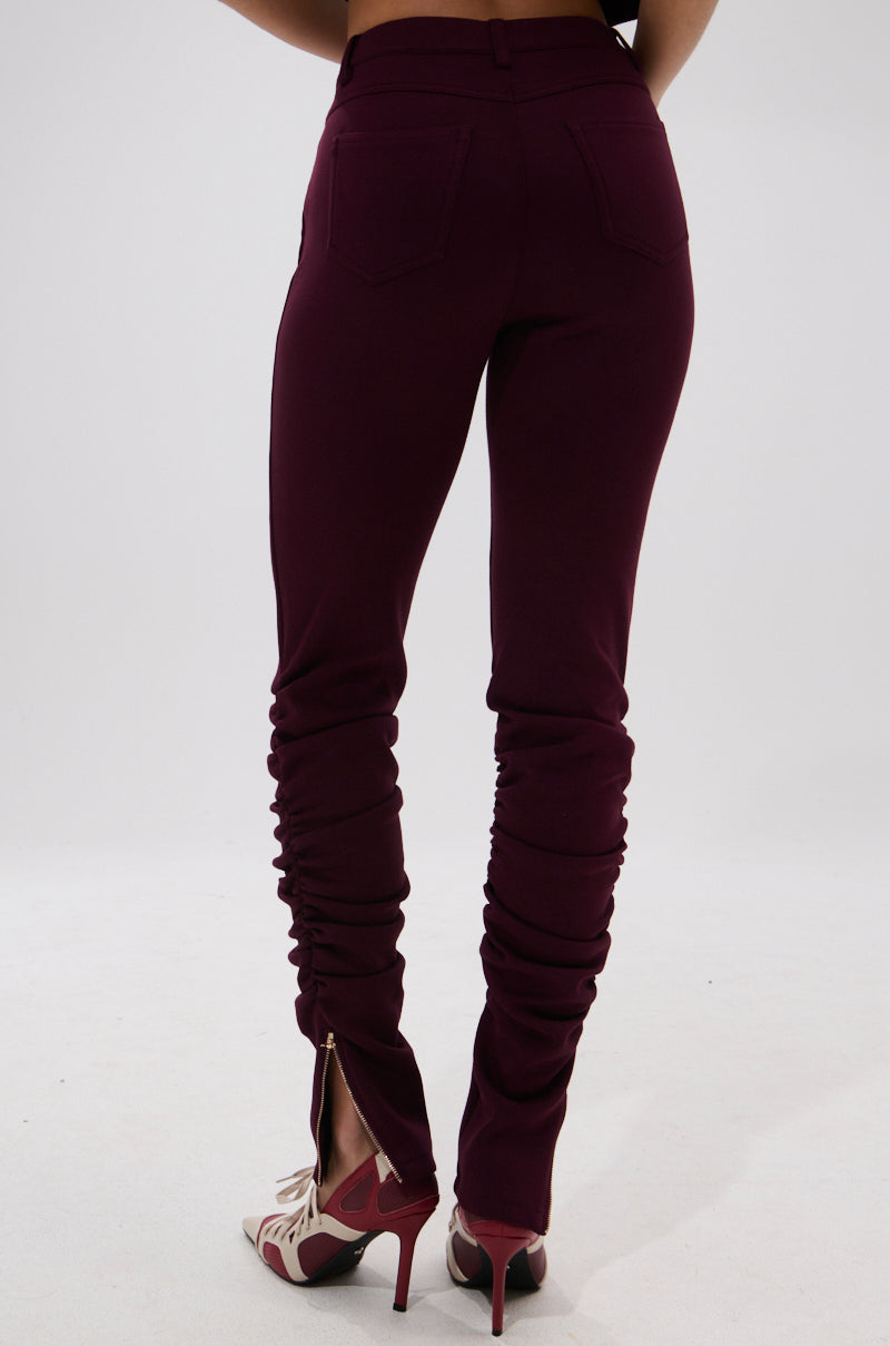 A person wearing the LYNX STACKED TROUSER IN BURGUNDY, featuring a high-rise skinny fit with back pockets and ankle zippers, paired with open-toe heels, stands against a plain white backdrop. 4