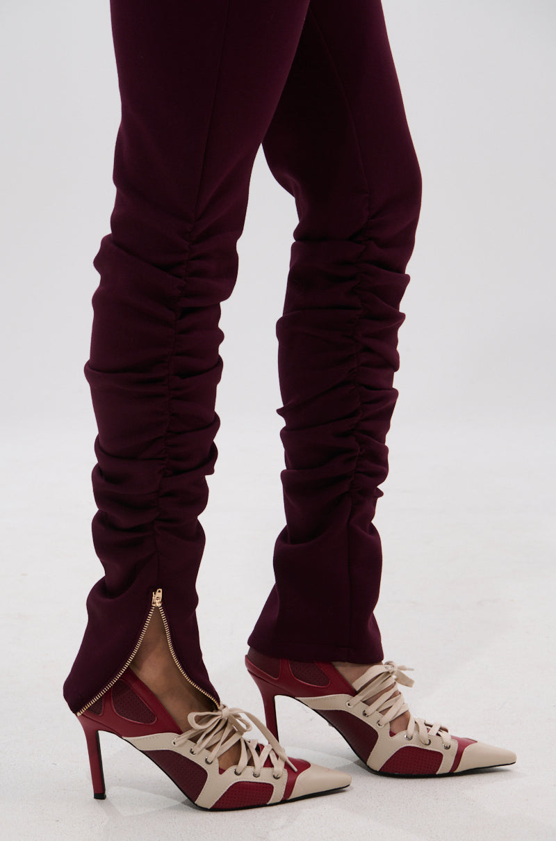 Wearing the LYNX STACKED TROUSER IN BURGUNDY featuring ankle zippers and ruching, paired with cream and burgundy lace-up high heels, a person stands on a white surface. 7