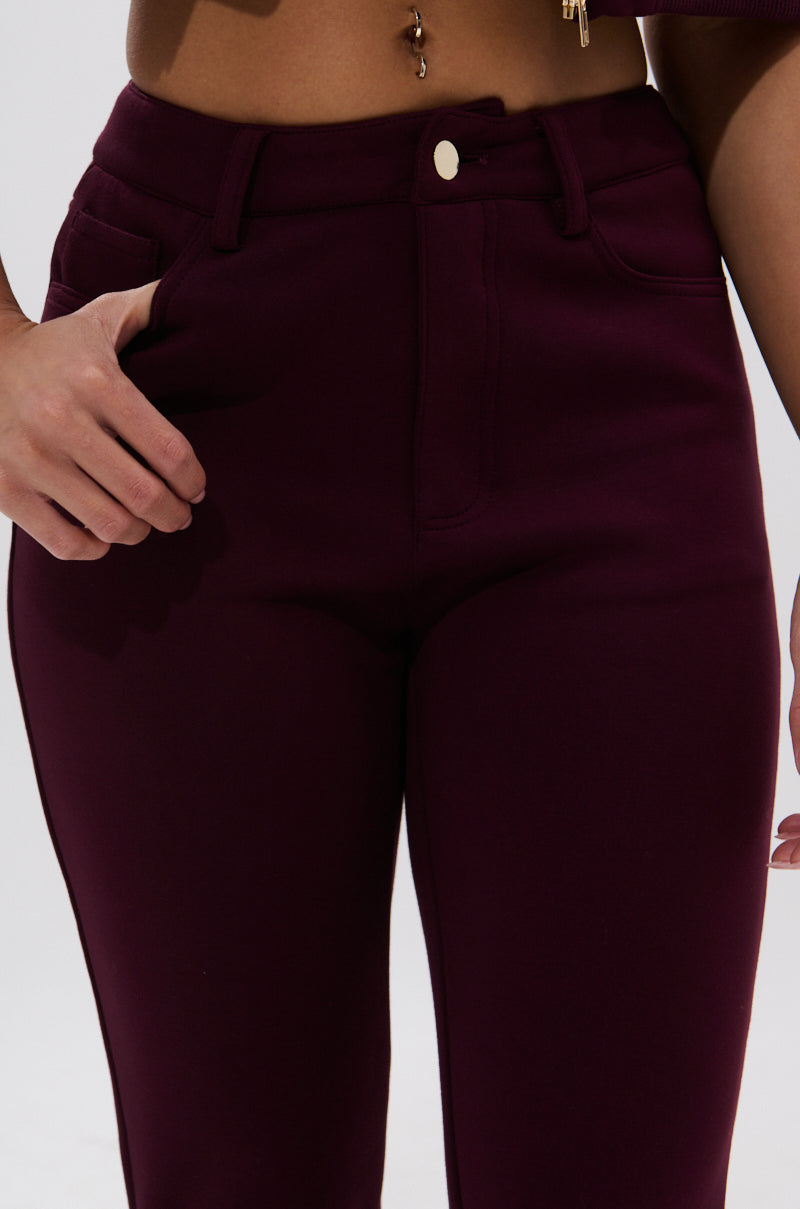 Wearing the LYNX STACKED TROUSER IN BURGUNDY, a person displays its high-waisted, skinny fit and gold button detail while resting a hand on the waistband; a belly button piercing is visible against a plain, light-colored background. 8
