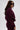 A woman with long dark hair, wearing large black sunglasses and the LYNX LAYERED BOMBER JACKET IN BURGUNDY, stands sideways and looks slightly toward the camera against a plain light background.