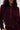 A woman with long brown hair and neutral makeup stands against a plain light background, wearing the LYNX LAYERED BOMBER JACKET IN BURGUNDY and matching pants. Her face is partially visible.