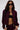 A woman with long dark hair stands against a plain background, hands in her pockets, wearing large black sunglasses and the LYNX LAYERED BOMBER JACKET IN BURGUNDY over a black crop top and maroon pants. Her belly button piercing is visible.