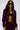 A woman with long brown hair models the LYNX Layered Bomber Jacket in Burgundy, styled with matching pants, black sunglasses, and a black crop top that shows her belly button piercing against a plain light background.