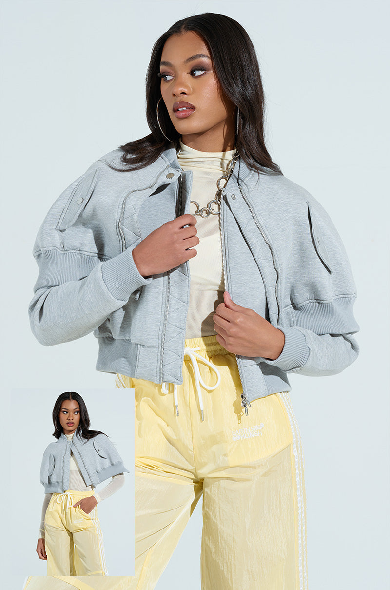 A woman strikes a pose in the LYNX Layered Bomber Jacket in Heather Grey, paired with yellow pants, clutching the collar. Her long hair cascades over hoop earrings and a chain necklace. A smaller image highlights her stylish and convertible ensemble in the bottom left corner.