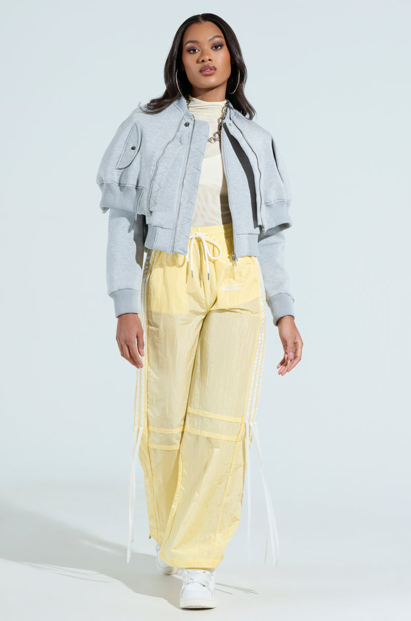 A person is posing against a light background, wearing the LYNX Layered Bomber Jacket in heather grey over a white top, with loose yellow drawstring pants. They have long, dark hair and are wearing white sneakers.