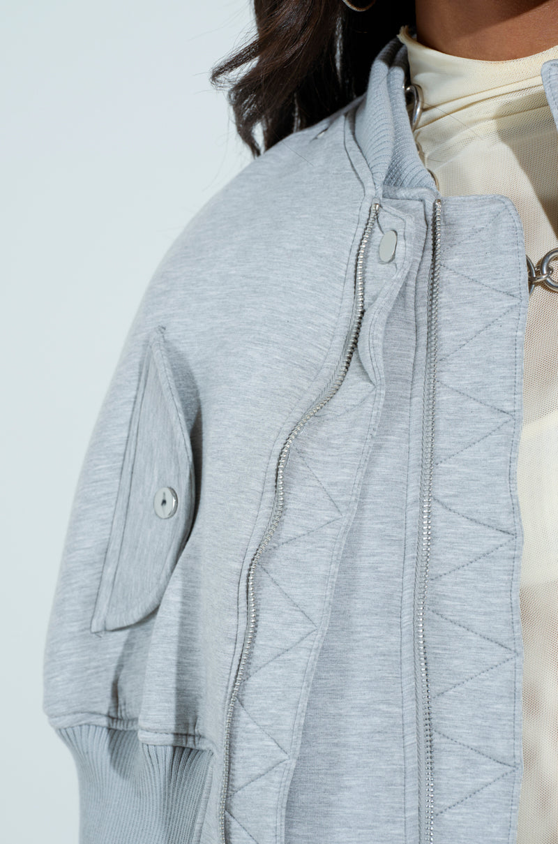 A close-up shows someone wearing the LYNX Layered Bomber Jacket in heather gray. Crafted from semi-stretchy scuba knit, it features a visible zipper and pocket detail. The cropped design hints at a beige top underneath, with only the torso and hair against a light backdrop visible.