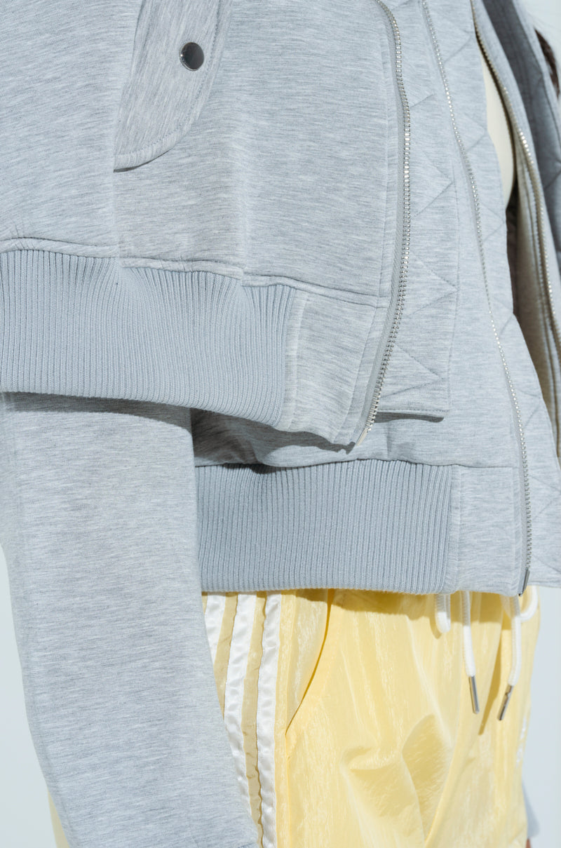 The image shows a close-up of a person wearing the LYNX Layered Bomber Jacket in heather grey, inspired by a classic bomber silhouette with ribbed cuffs. The jacket is paired with shiny yellow pants featuring white side stripes.