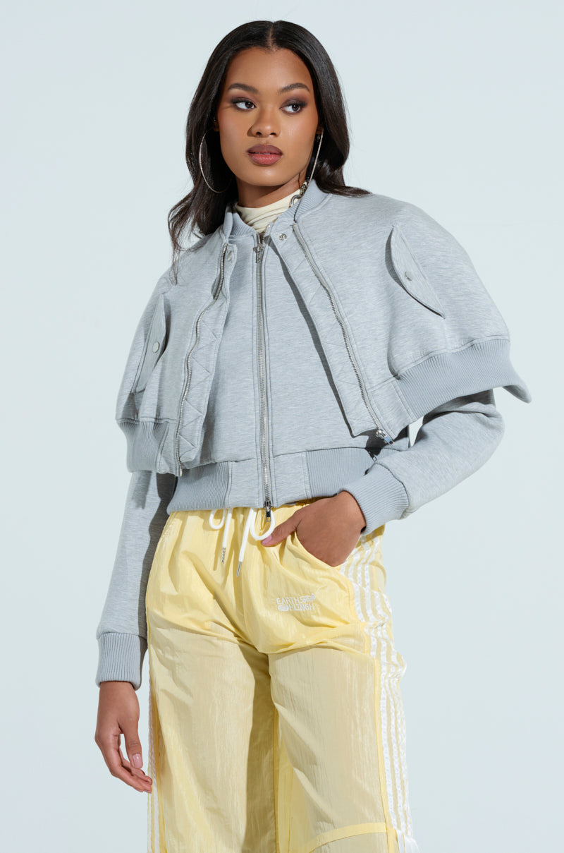 A woman poses confidently against a plain light background, wearing the LYNX Layered Bomber Jacket in heather grey, featuring unique layering and drawstring details, paired with light yellow Adidas track pants.