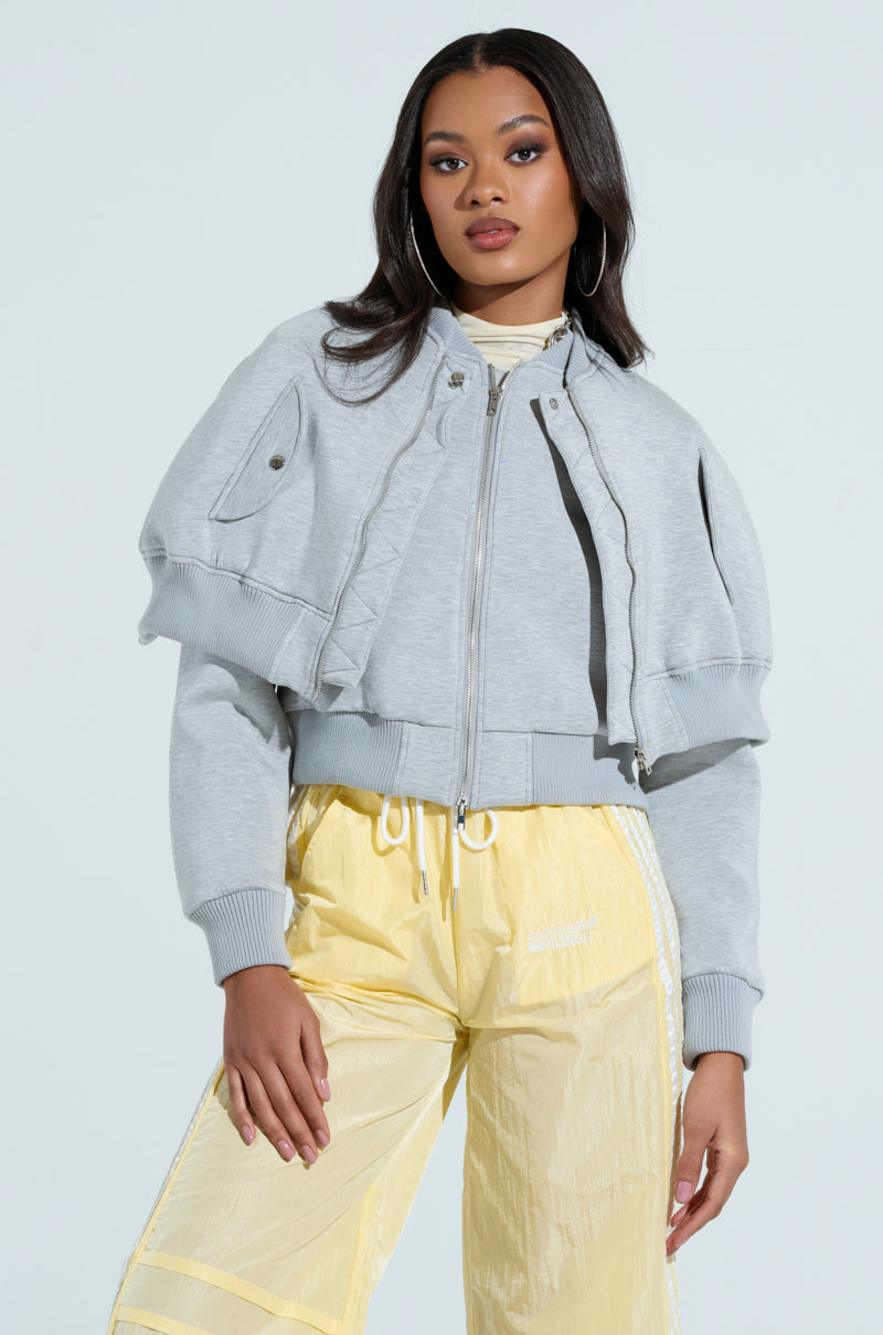 Someone in the LYNX Layered Bomber Jacket in Heather Grey and yellow drawstring pants, showcasing modern design with layered elements. They stand against a plain light background, sporting long hair and embodying effortless style.