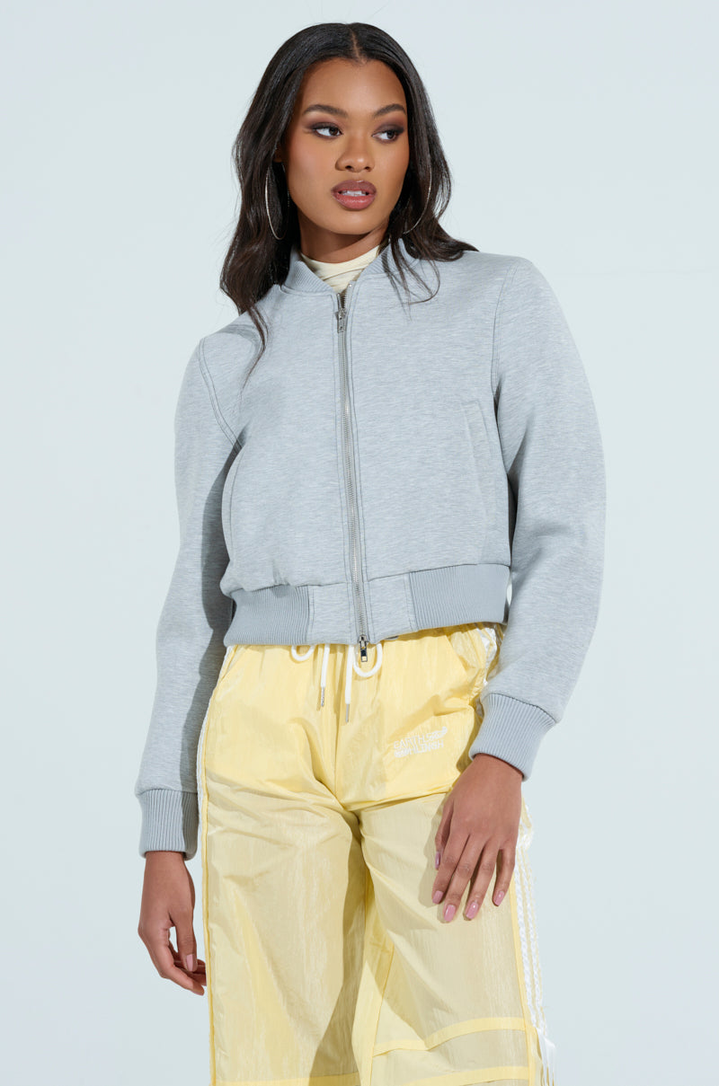 Wearing a LYNX Layered Bomber Jacket in heather grey and translucent yellow pants, a person with long hair stands against a plain background, looking slightly to the side.
