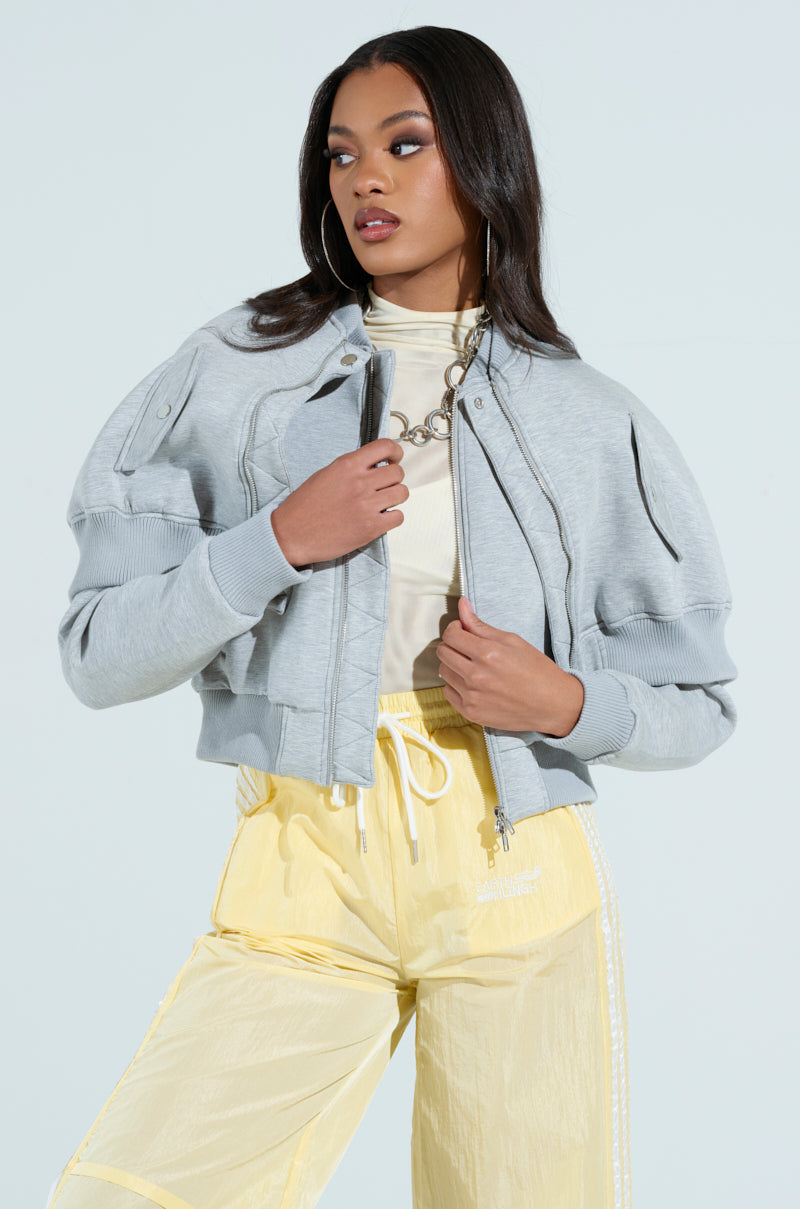 A woman in the LYNX Layered Bomber Jacket in heather grey over a white top stands against a plain background. She pairs it with yellow pants, holding the jacket collars confidently. Her long hair flows freely, accessorized with hoop earrings and a silver chain necklace.