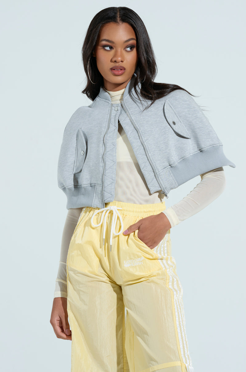 The woman wears a LYNX Layered Bomber Jacket in heather grey over a sheer white long-sleeve shirt and light yellow Adidas pants. Positioned against a light blue backdrop, she gazes sideways with a neutral expression.