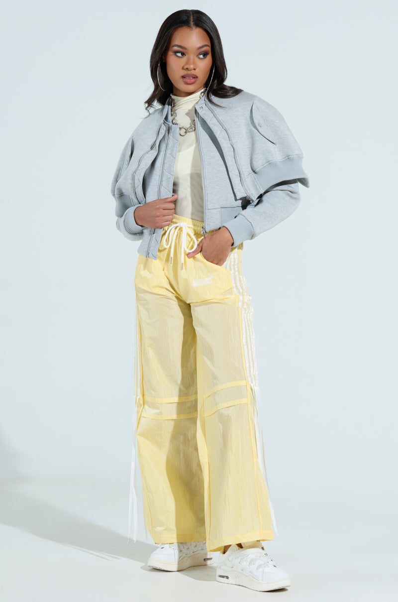 A person poses confidently in the LYNX Layered Bomber Jacket in heather grey and a light yellow turtleneck, paired with baggy yellow pants with white stripes. White sneakers complete the look, highlighted against a plain, light background to showcase the semi-stretchy scuba knit fabric.