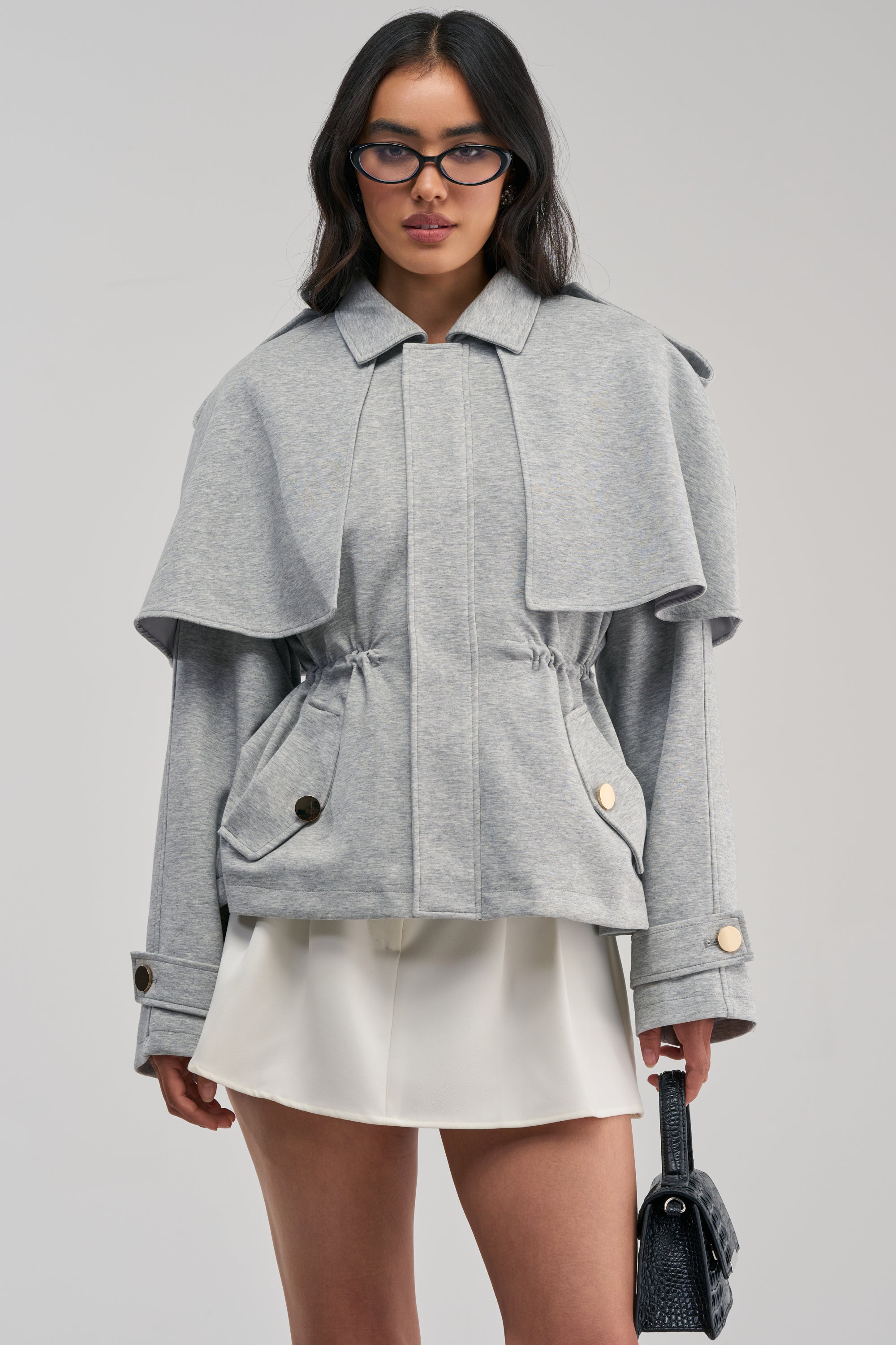 A woman wearing glasses stands against a plain background, dressed in the LONDON TOWN DOUBLE LAYER JACKET in light gray and a white skirt, while holding a small black handbag.