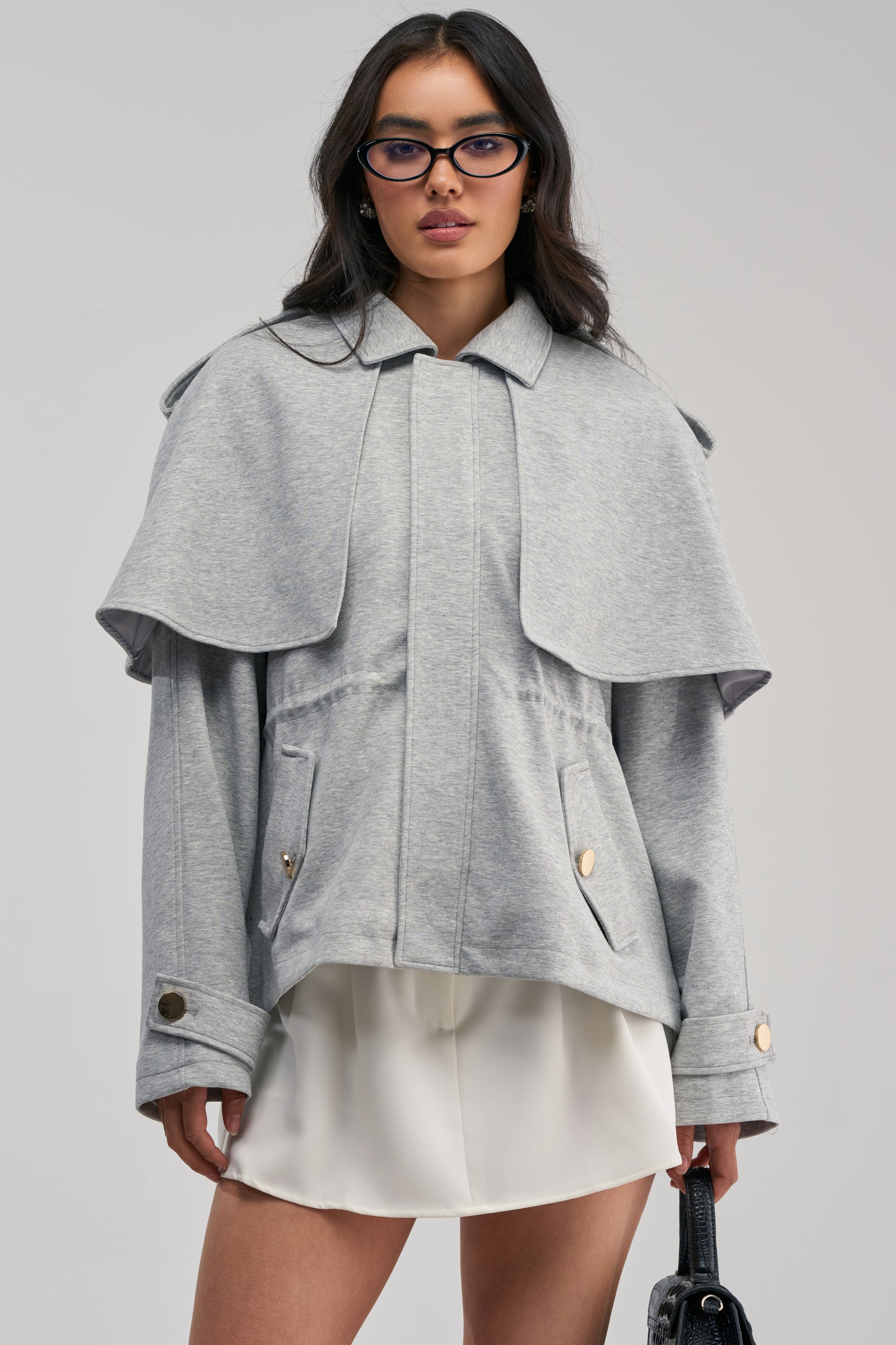 Wearing the LONDON TOWN DOUBLE LAYER JACKET in light gray over a white skirt, a woman accessorized with glasses and carrying a black handbag poses against a plain background.