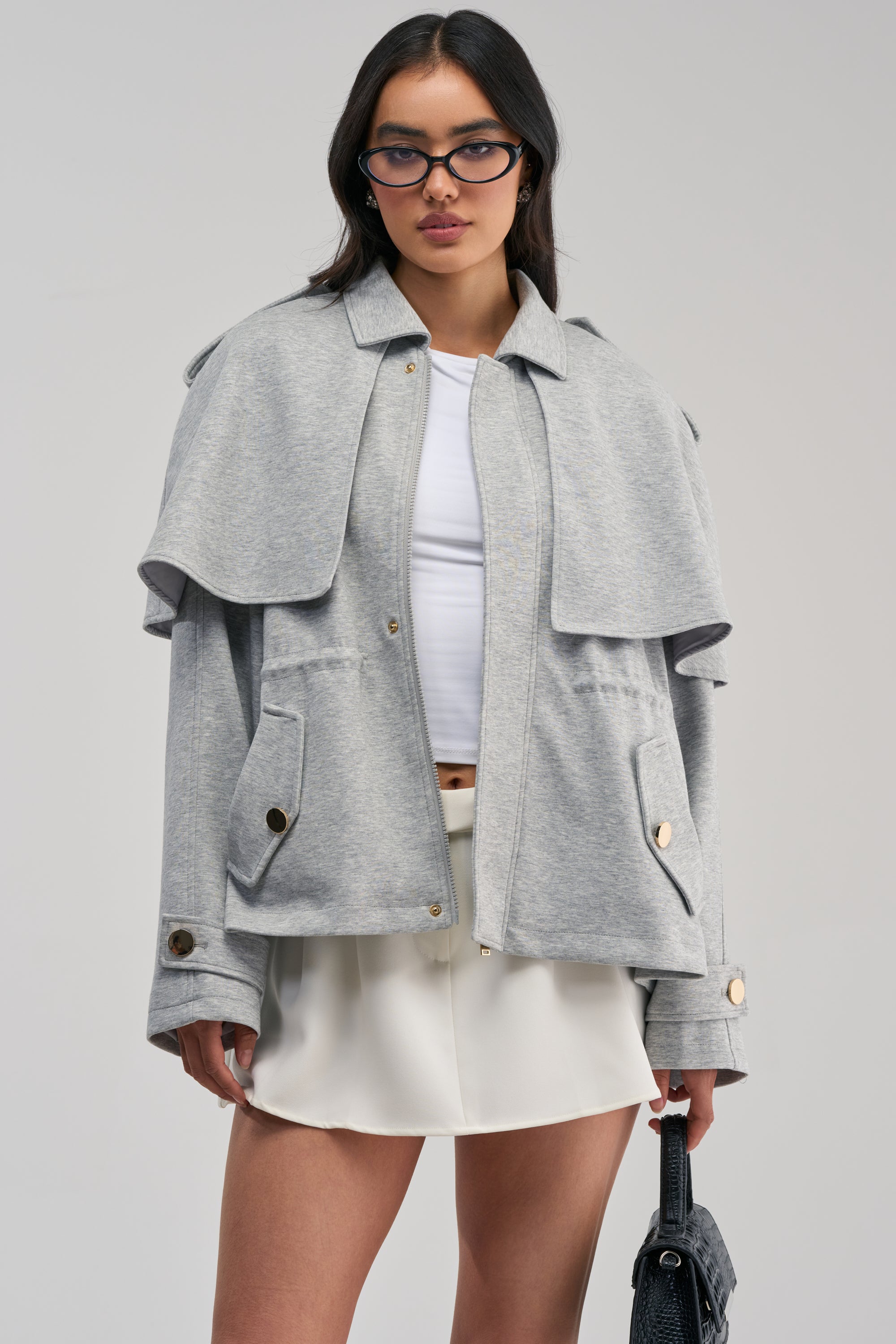 A woman stands against a plain background wearing the LONDON TOWN DOUBLE LAYER JACKET in light gray with layered shoulders, paired with a white top and short white skirt, and holding a black bag.