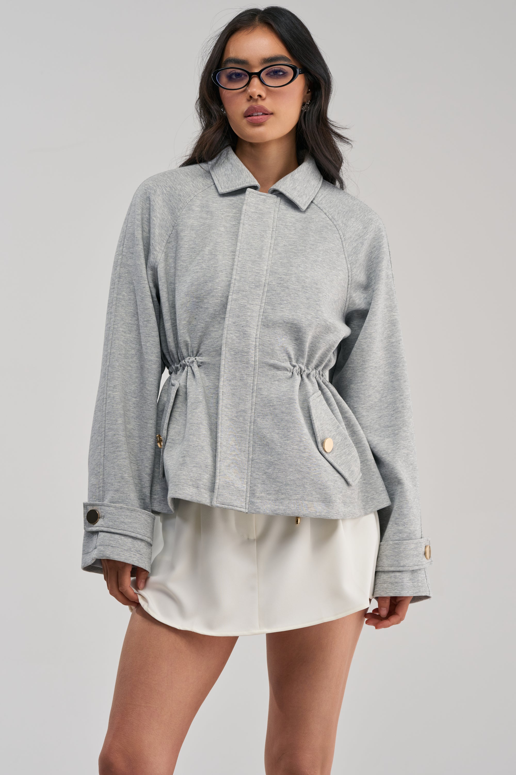 A woman with long dark hair and glasses stands against a plain light background, wearing the LONDON TOWN DOUBLE LAYER JACKET—a grey scuba-knit style featuring a drawstring waist and gold buttons—paired with a white mini skirt.