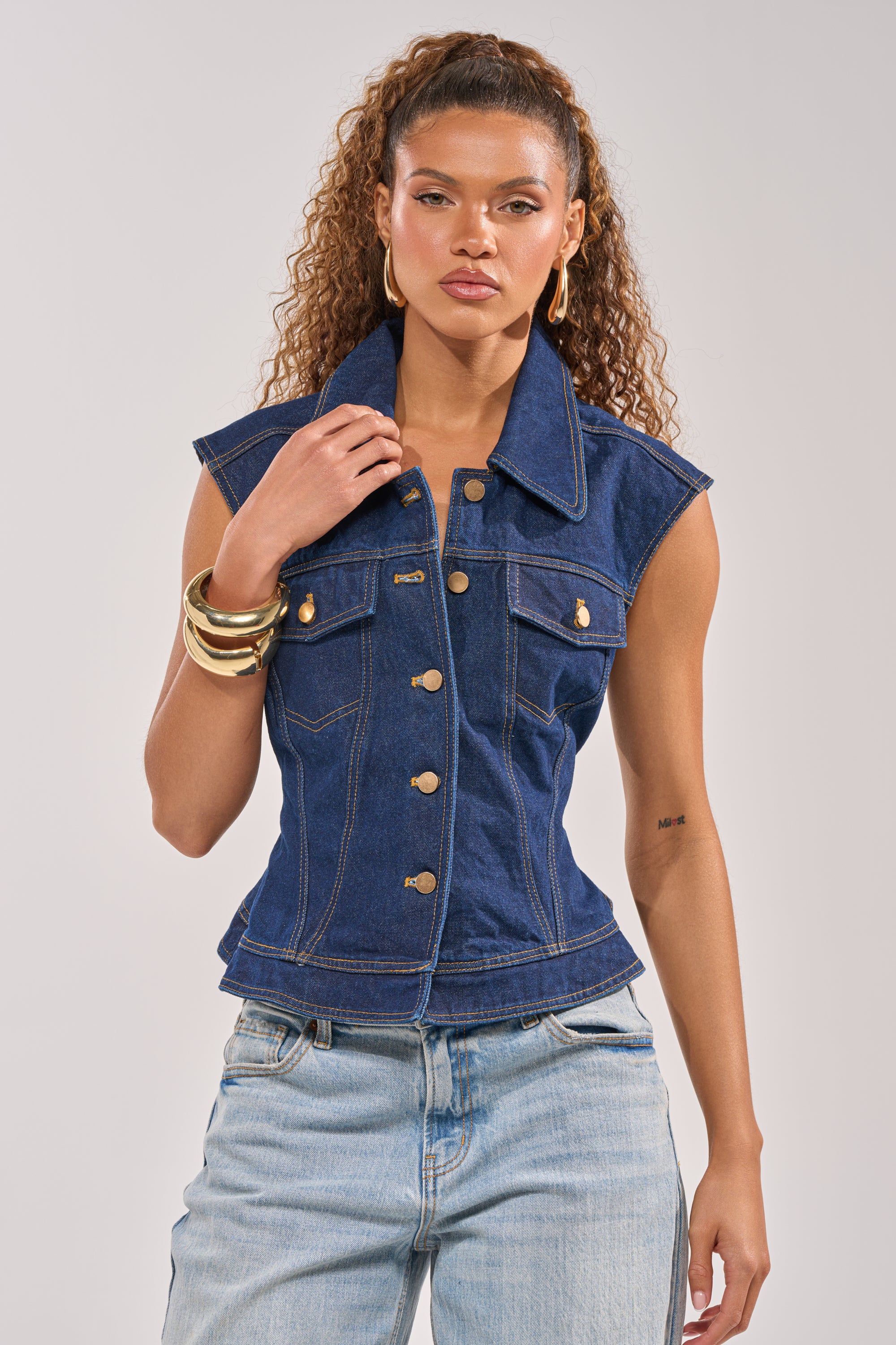 A woman with curly hair exudes sophistication in large hoop earrings, gold bangles, the BIRDIE DENIM VEST with gold buttons, and light blue jeans as she poses confidently against a plain background.